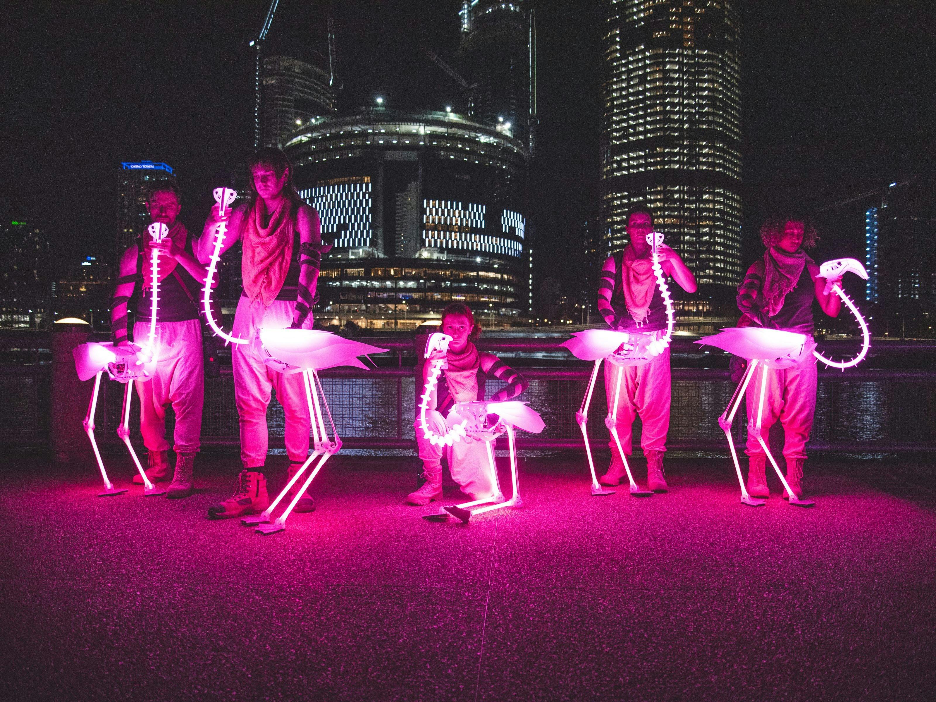 A group of 5 people all holding light-up flamingo puppets in front of the Brisbane river