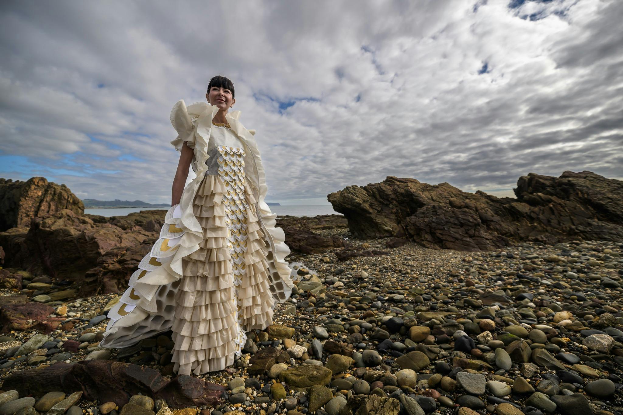 Model in flowing cream and gold dress stands on a rocky beach with dramatic sky