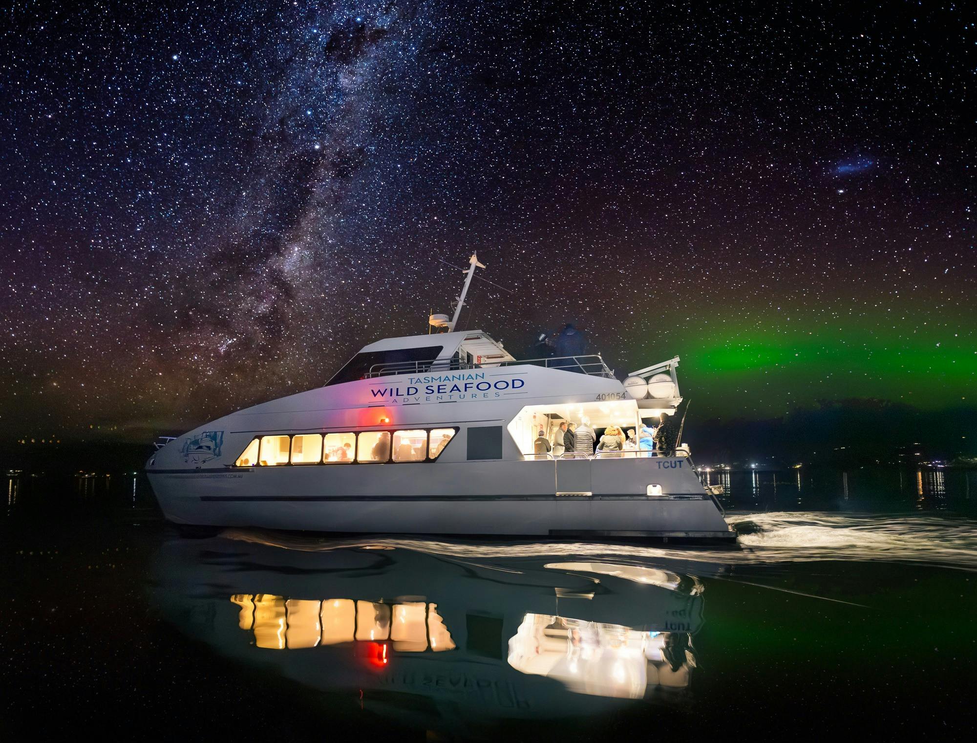 A 2 storey catamaran glides along the water at night with the aurora australis in the background