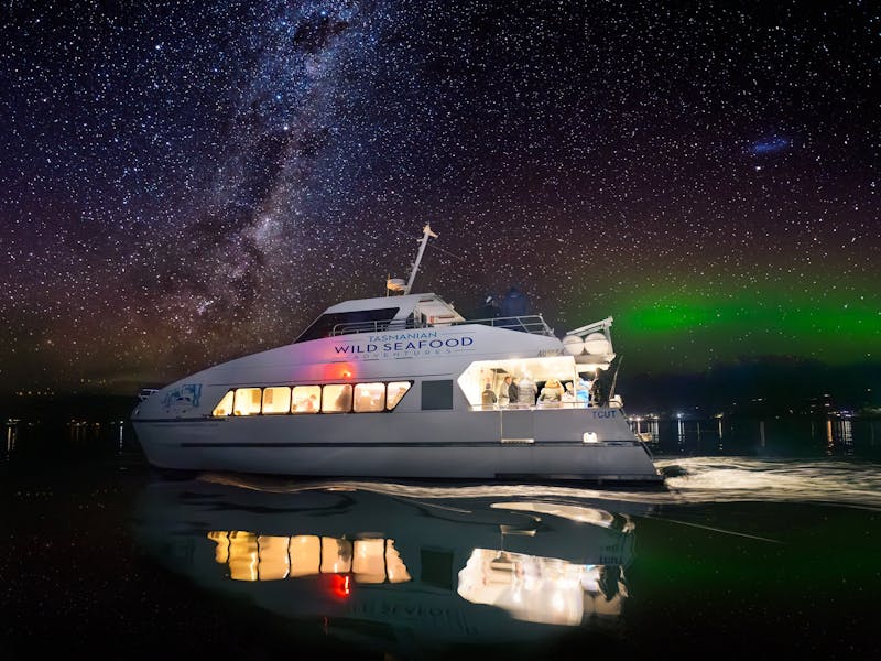 A 2 storey catamaran glides along the water at night with the aurora australis in the background