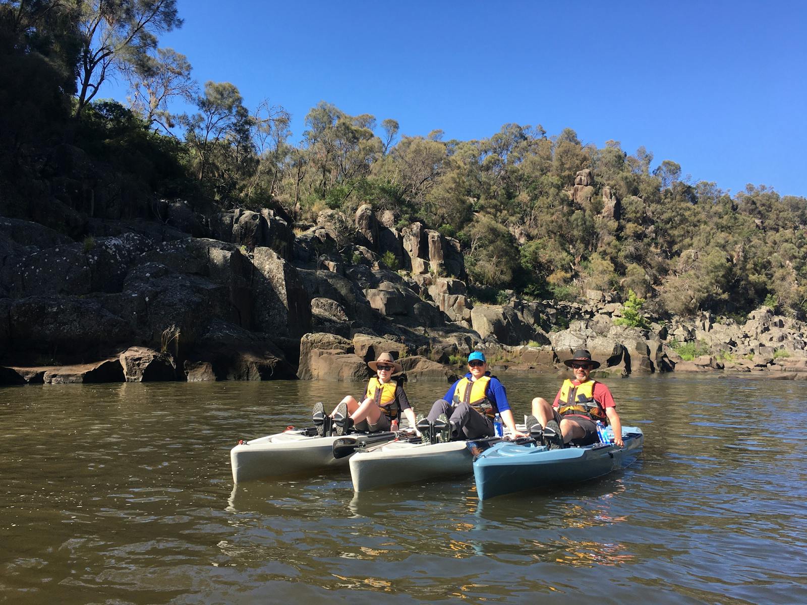 3 people each on  kayak with scenic cliff and bushland in the back ground