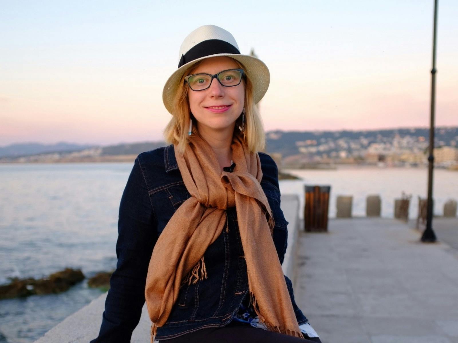 Woman in hat and scarf smiles by waterfront at sunset.