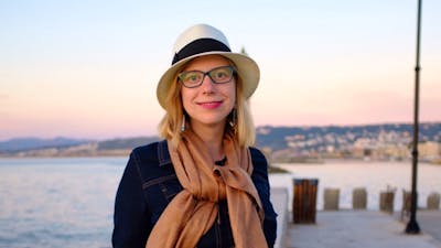 Woman in hat and scarf smiles by waterfront at sunset.