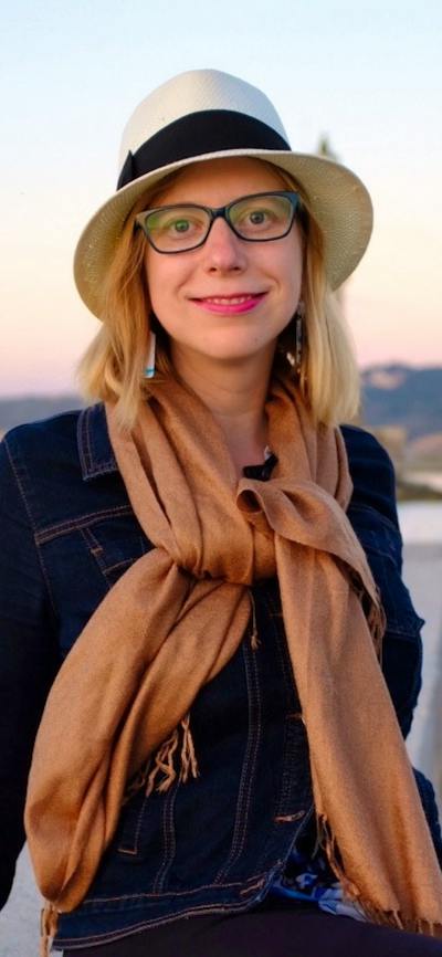 Woman in hat and scarf smiles by waterfront at sunset.
