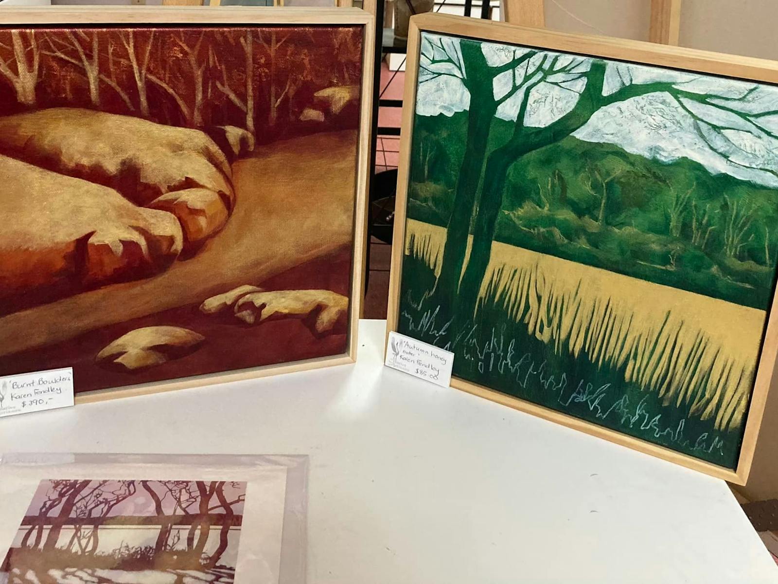 two paintings. One of trees and paddock in green tones and one of rocks and trees in browny orange
