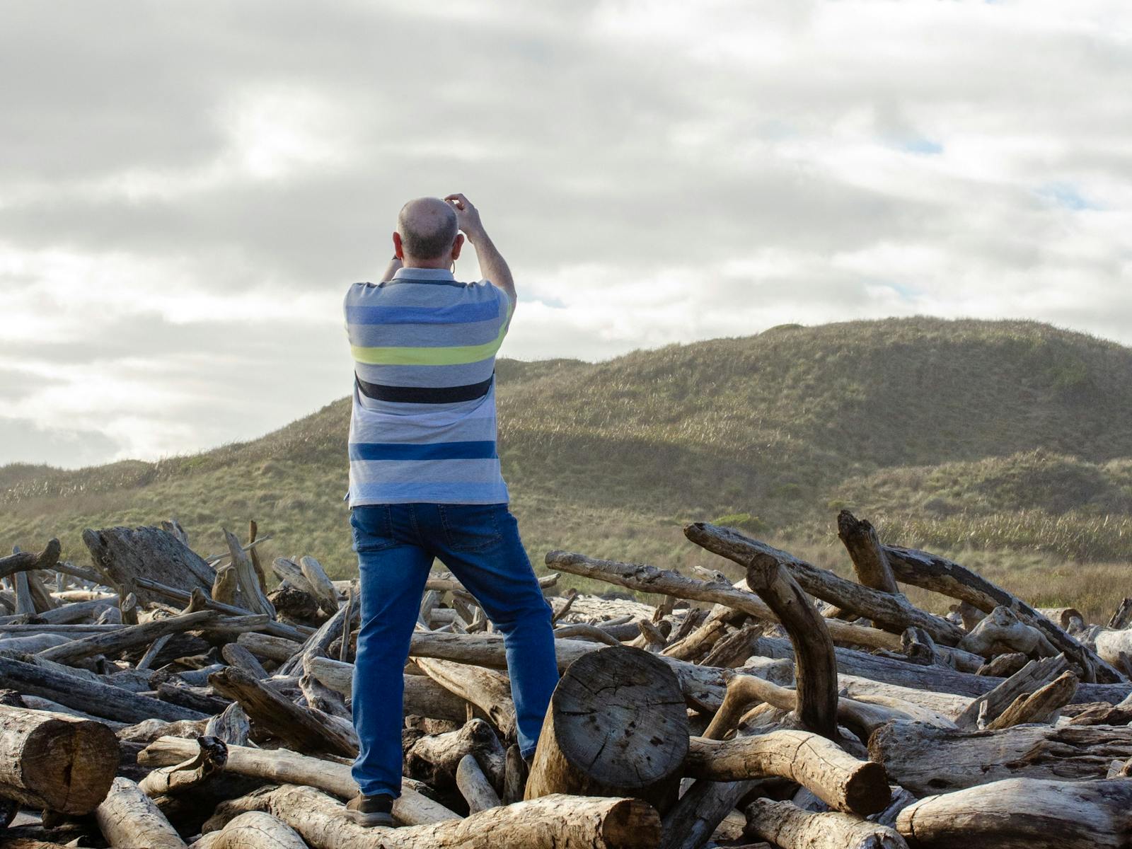Man standing on large driftwood taking a photo of the scene of drift wood and hills in front of him
