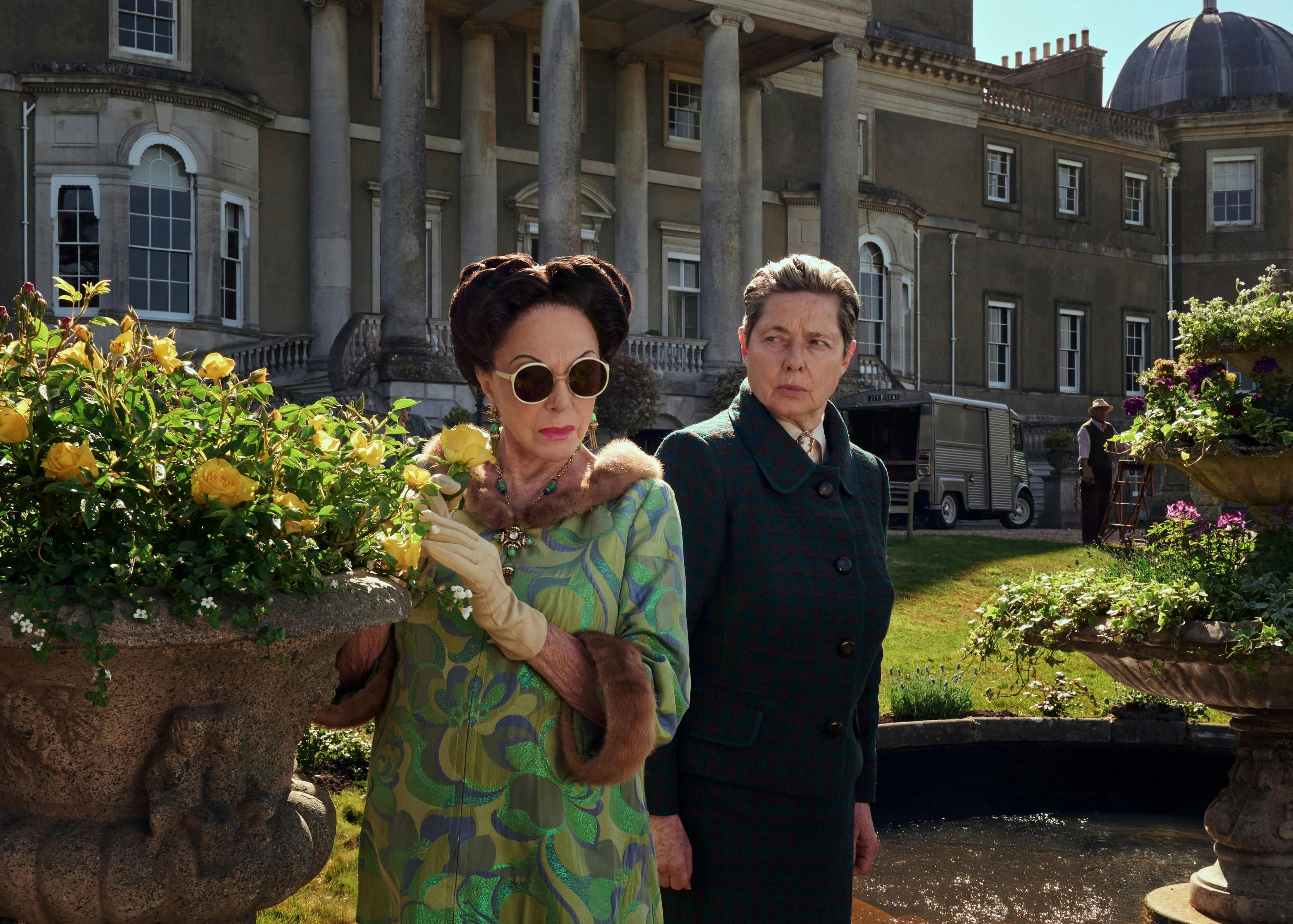 Two women stand in manor house garden next to yellow roses