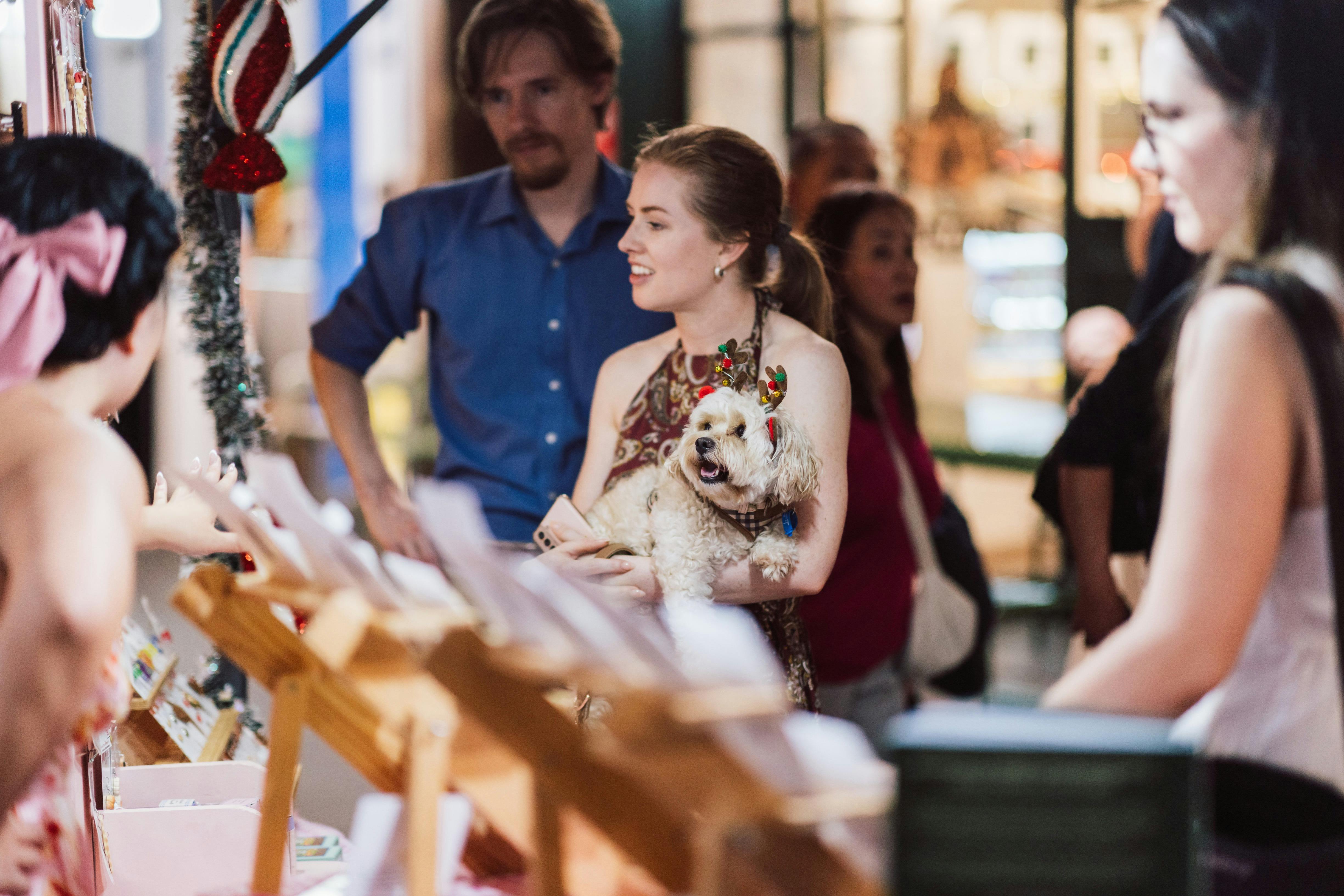 Woman holds a small dog wearing festive reindeer antlers while browsing a Christmas market stall.
