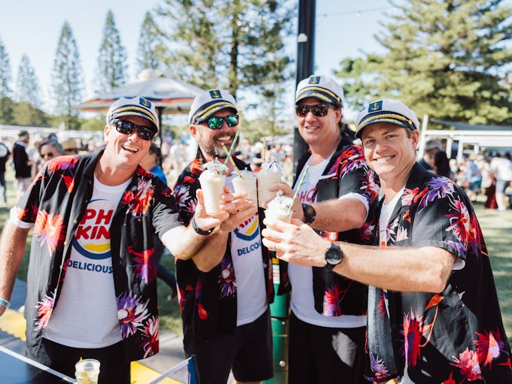 4 men cheersing their drinks towards the camera, wearing matching black and red Hawaiian shirts