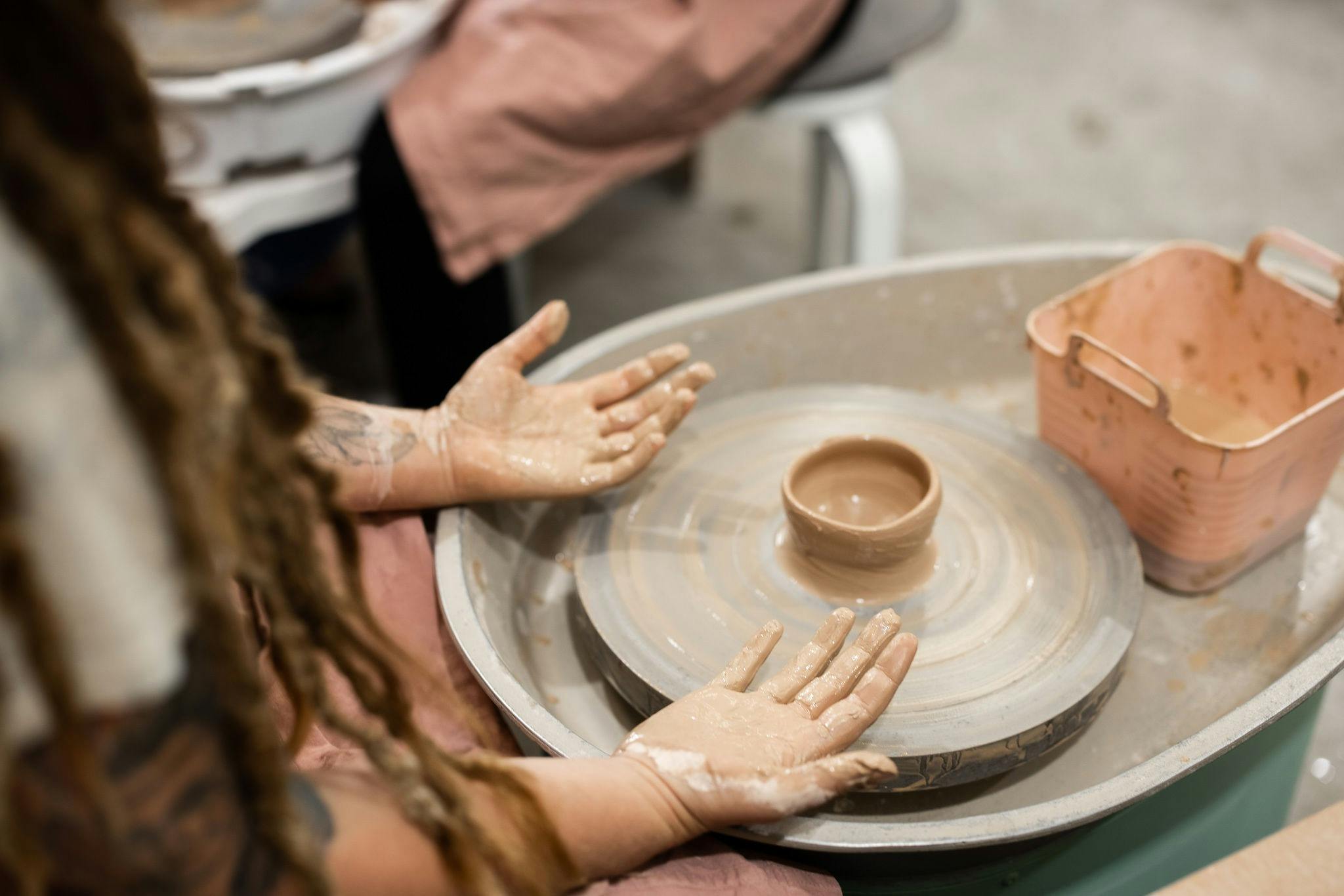 Picture shows a person seated at a pottery wheel with a small clay pot and dirty hands