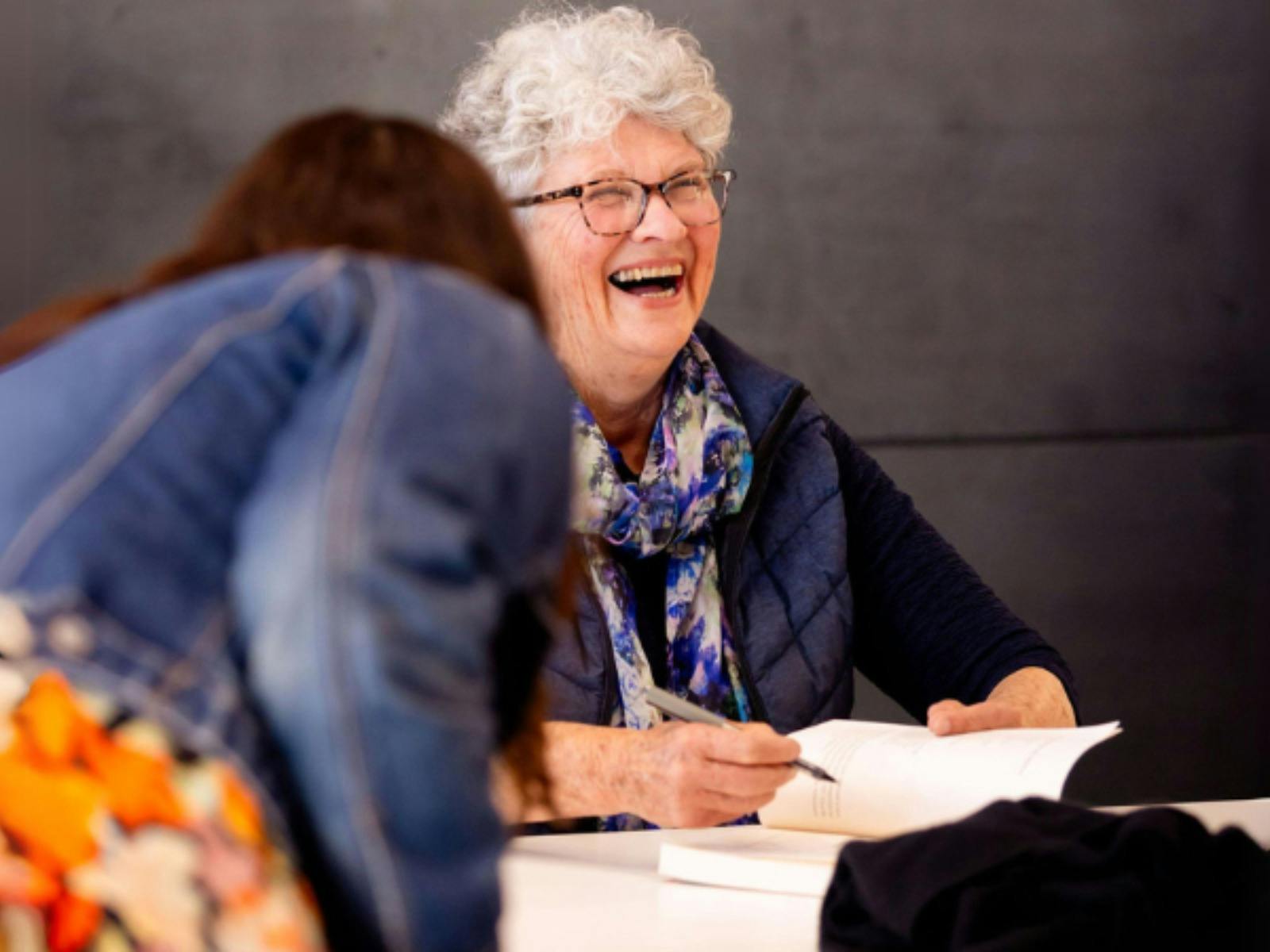 A smiling woman with short curly hair laughing while signing a book for a festival visitor.