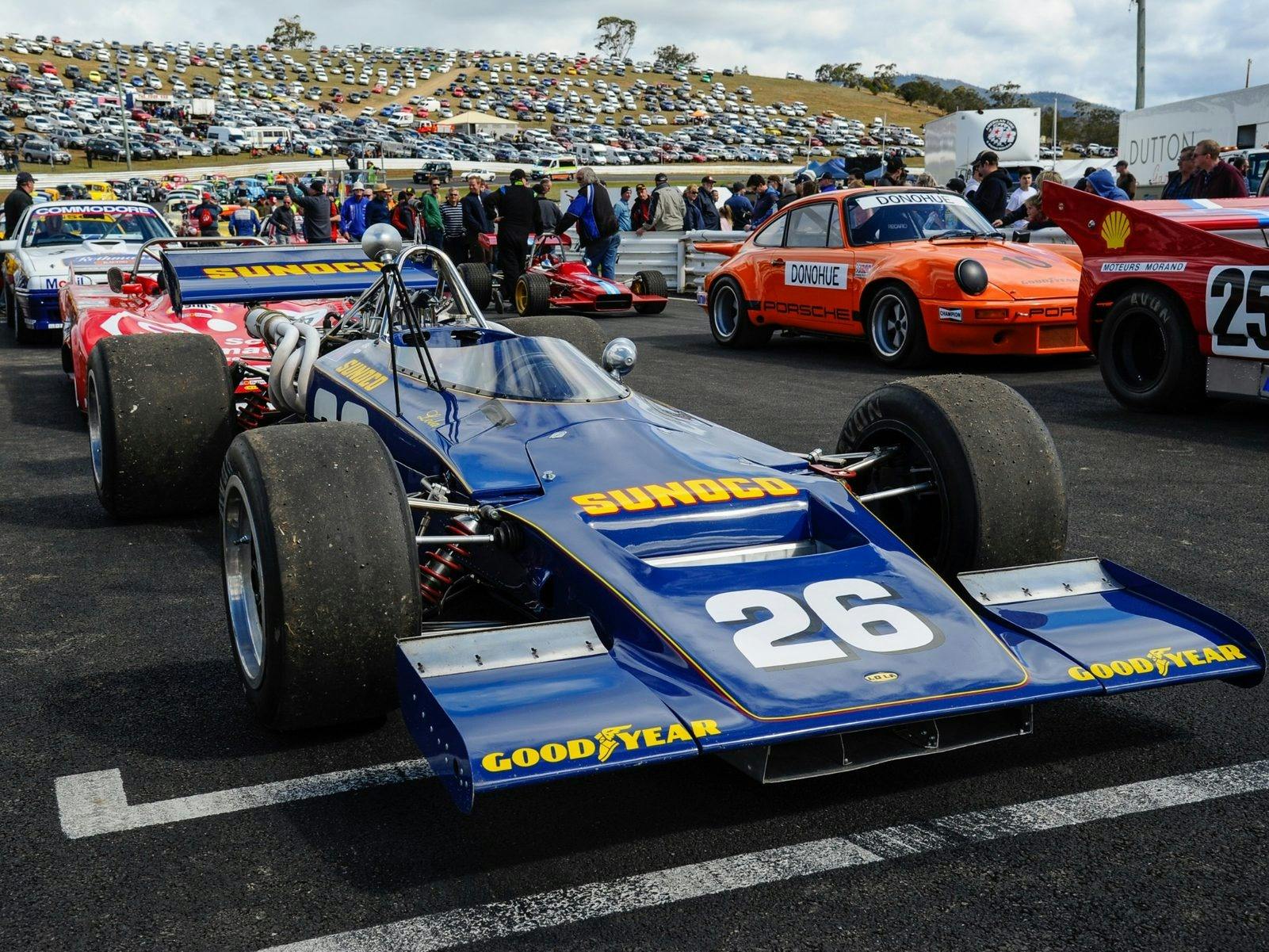 Blue open wheeler race car with number 26 on it, on the start grid with people walking past