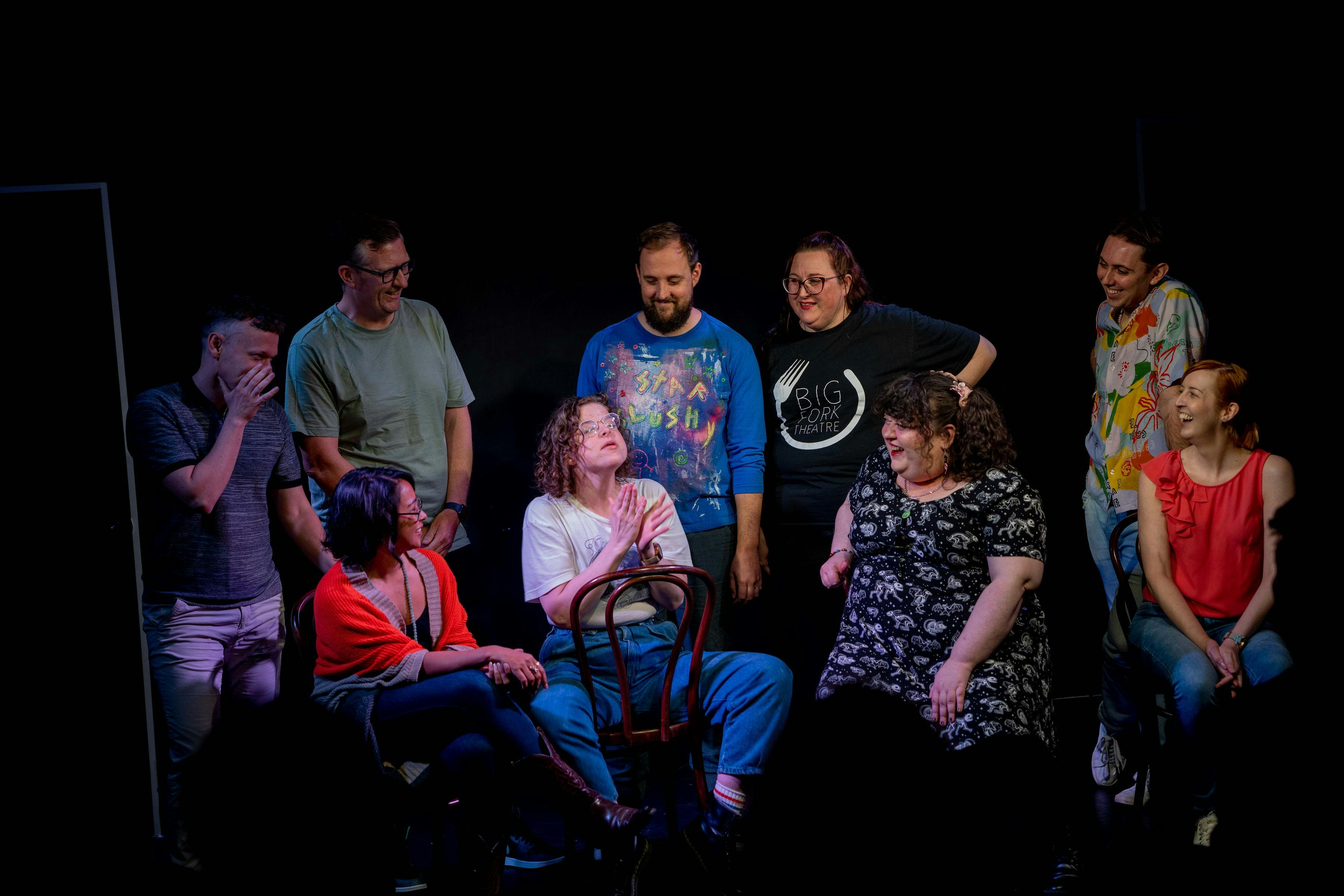 four performers seated and five stand behind them looking on as the middle performer tells a tale
