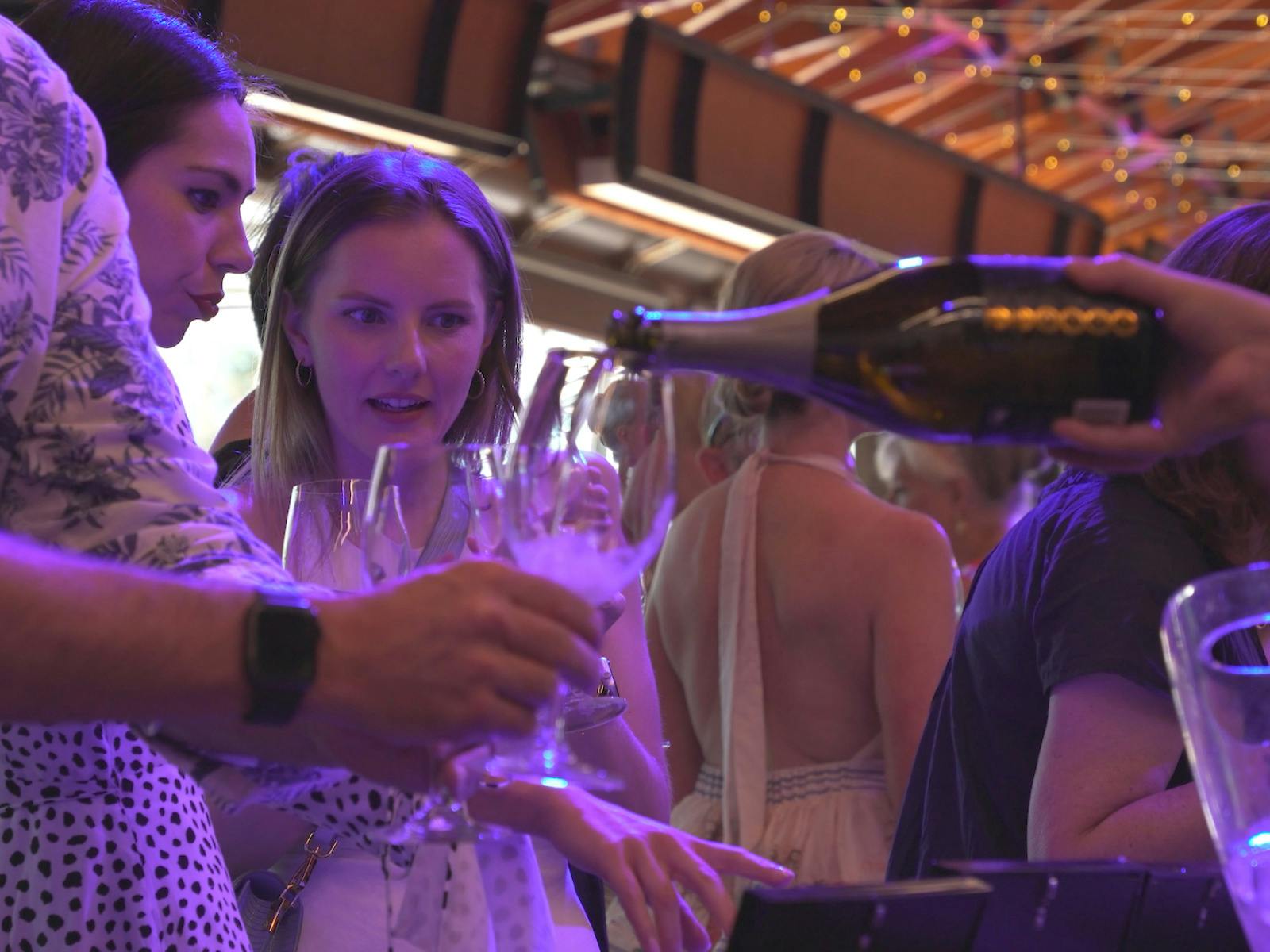A server pours sparkling wine into guests’ glasses as two women watch, bathed in purple light