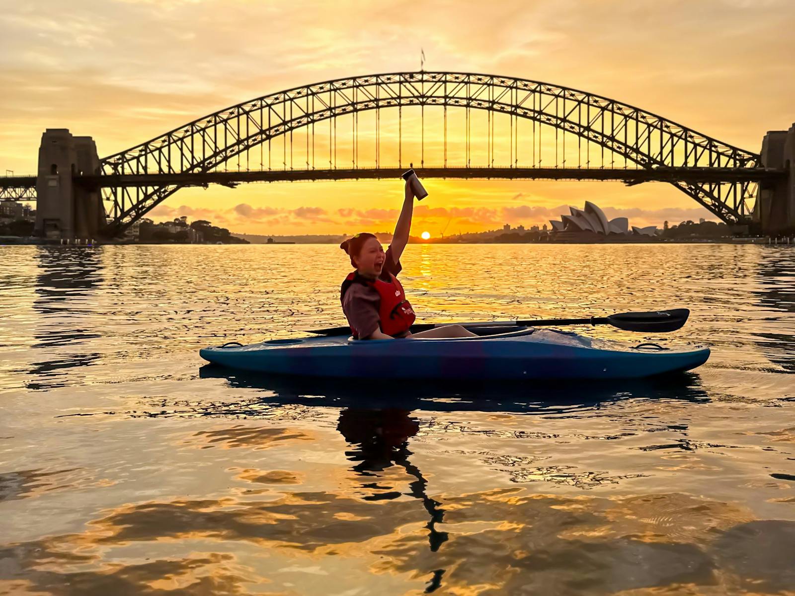 Lady cheering her coffee cup in the air from her kayak as sun rises over sydney harbour