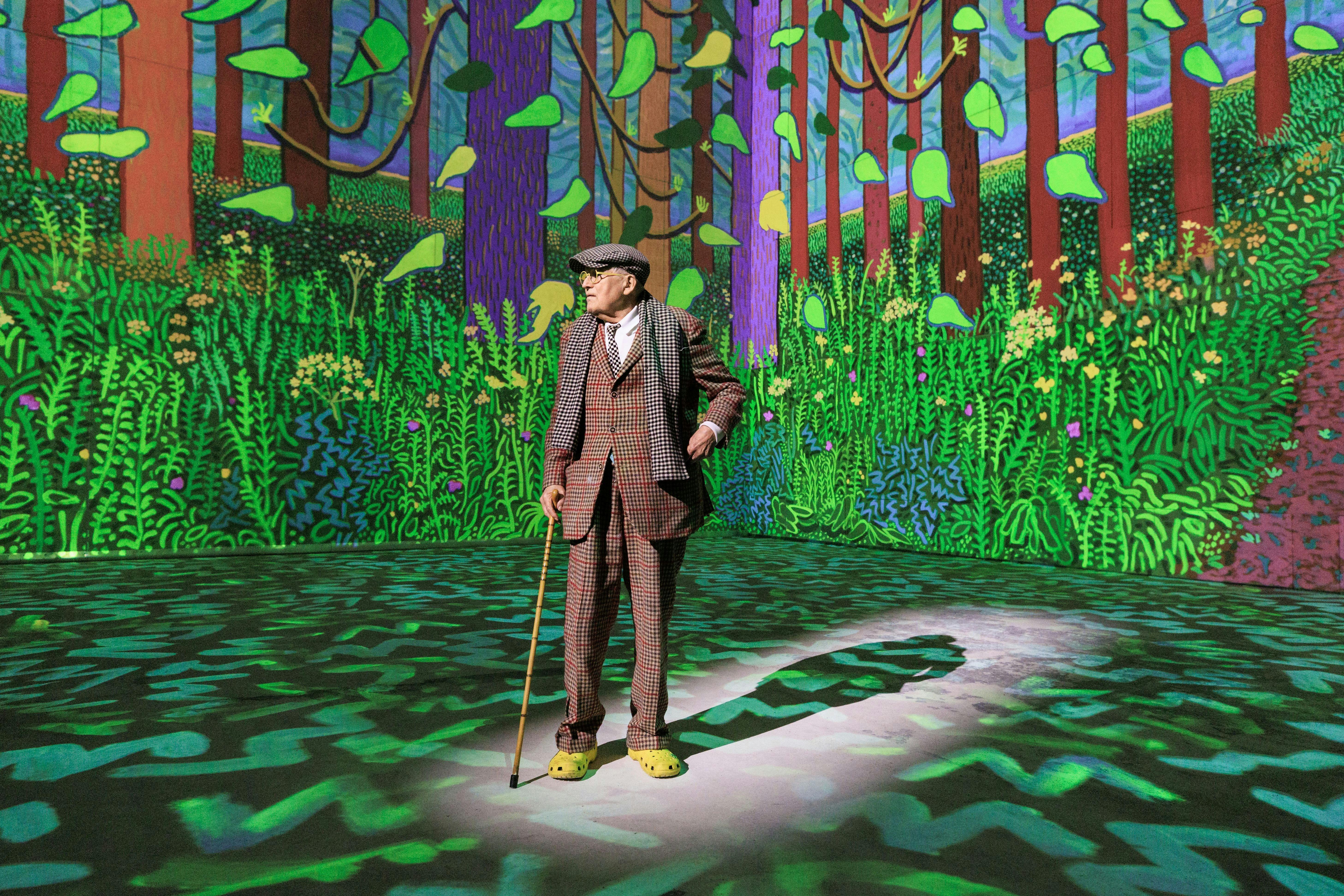 An elderly man in a plaid suit and flat cap stands with a cane on an immersive art display.