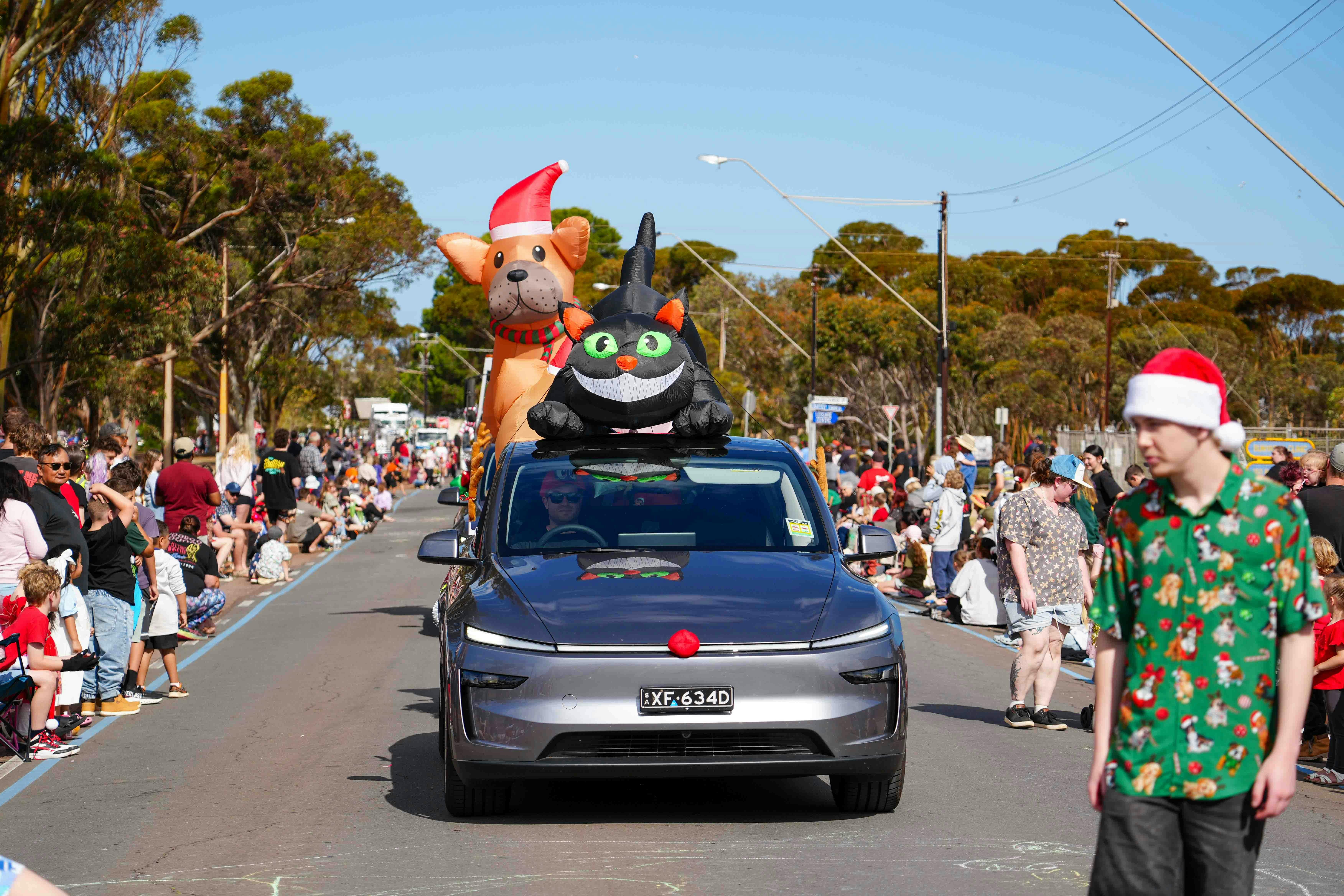 A grey tesla on the road with large inflatable cat and dog in Christmas hats tied to the roof