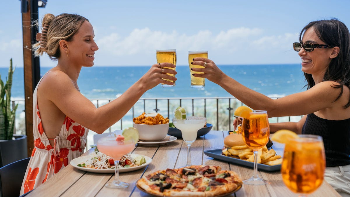 Two women toasting with beer over a table of food dishes
