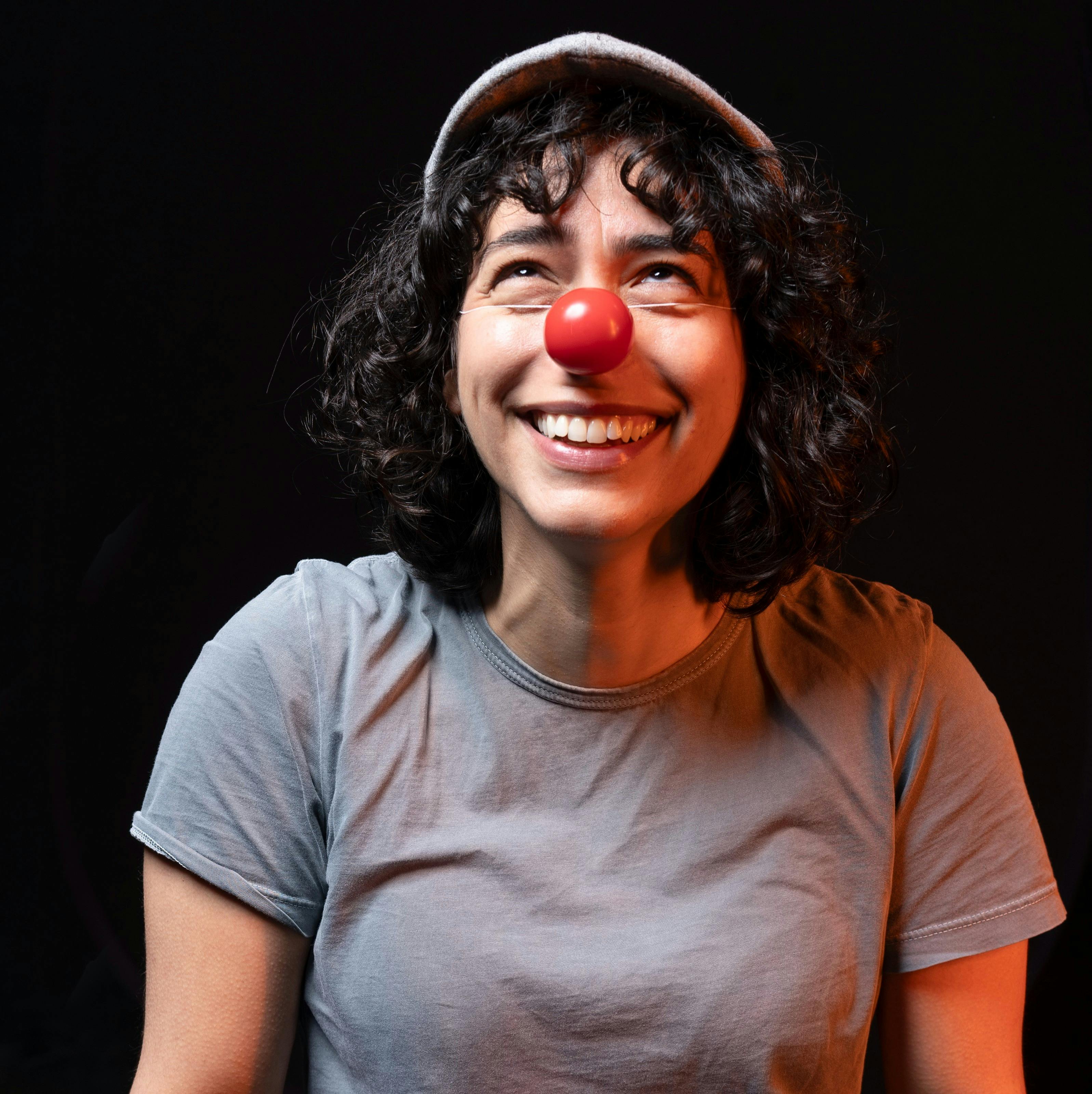 A woman wearing a clown nose stands against a black background.