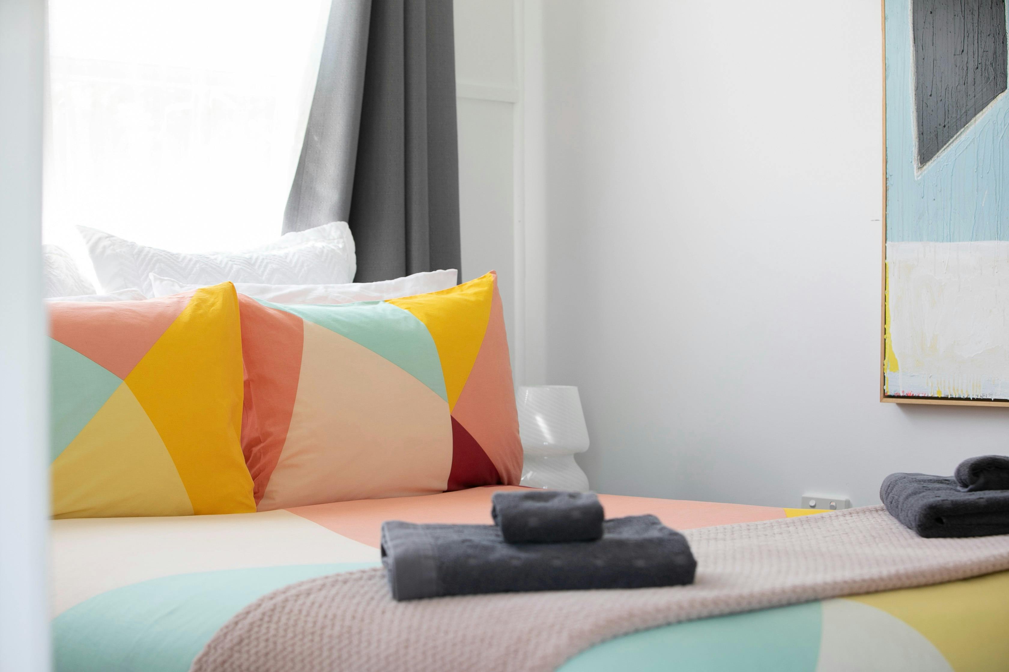 A queen sized bed with brightly cloured linen and a folded towel with a pink and blueainting.