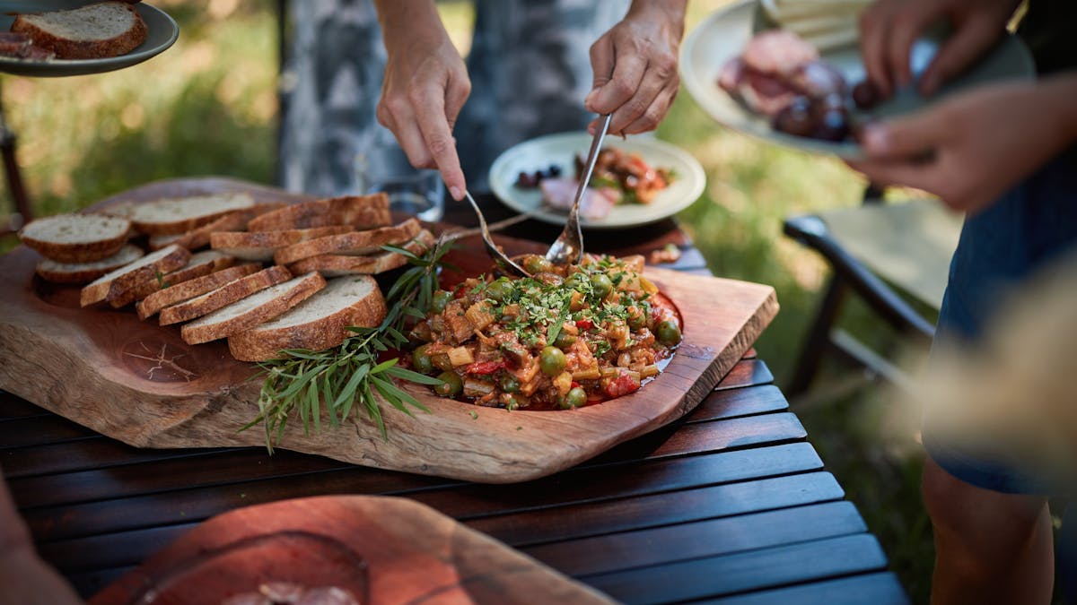 Bread and a vegetable dish on a a wooden board with someone serving themselves.