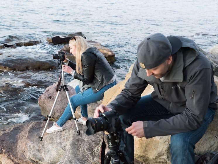 Man and women on rocks waters edge with camera on tripod