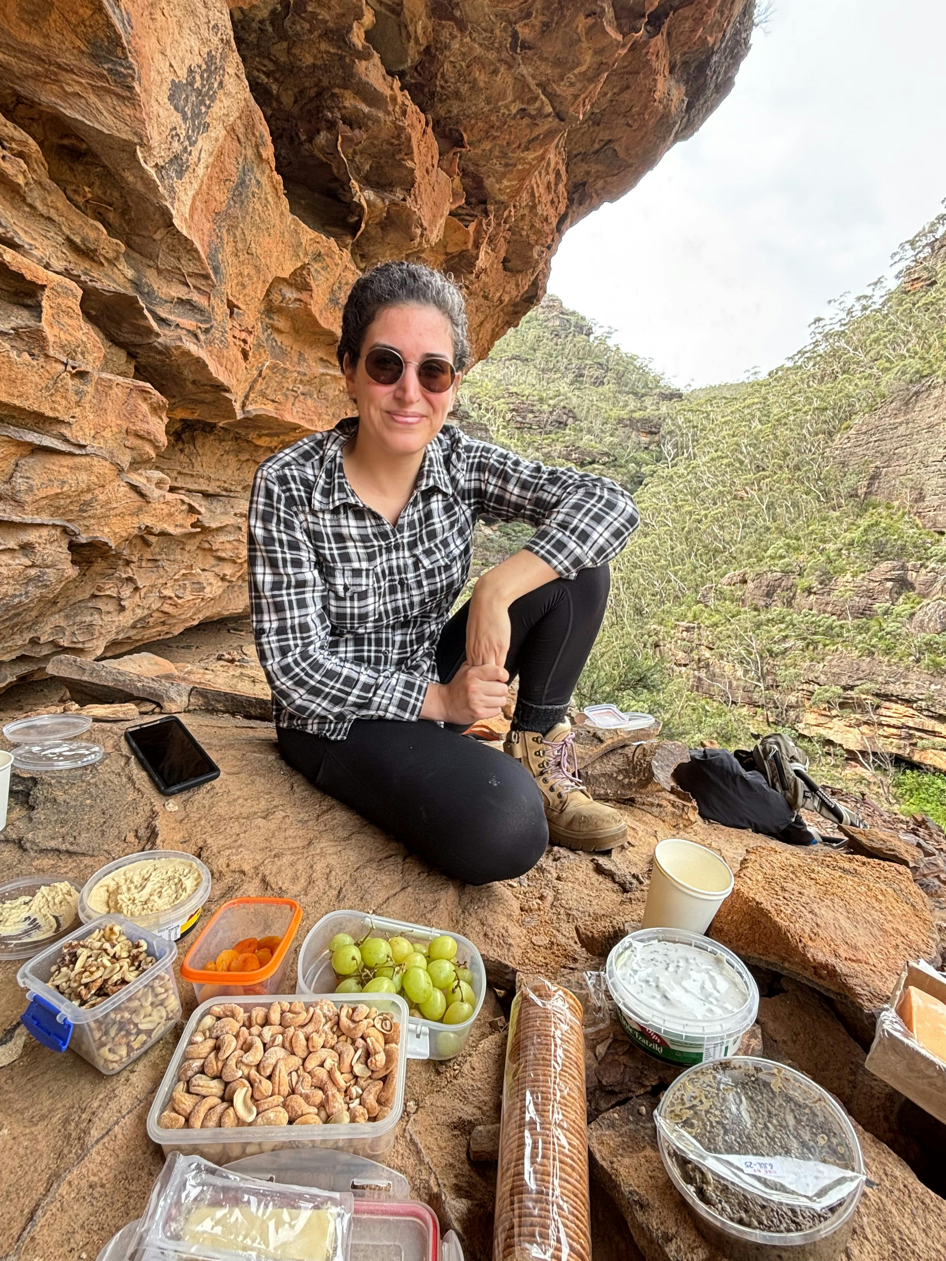 A woman wearing a checked shirt sitting down in front of a picnic lunch near a sandstone cliff.