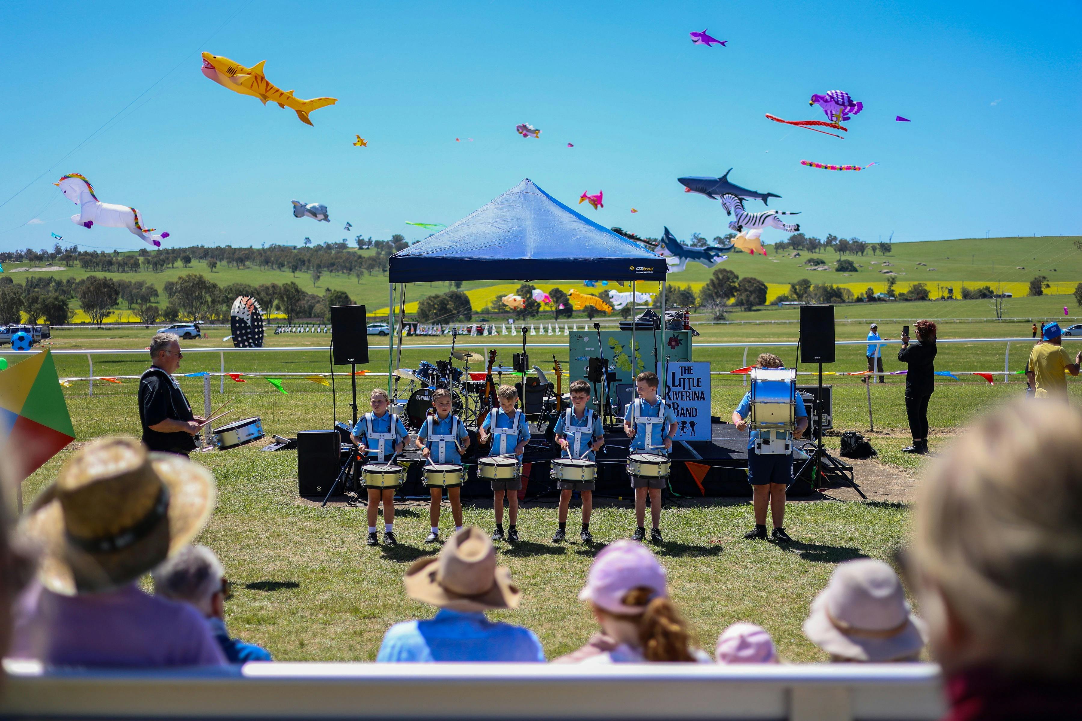 An audience watching a main stage music performance by a local school with kites flying in the back.