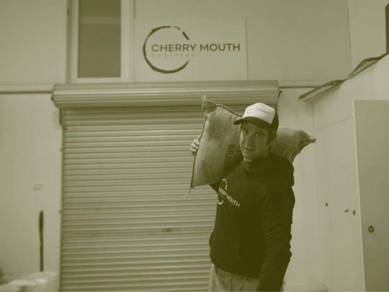 Man outside roller door with sack perched on shoulder and Cherry Mouth coffee wall sign above head