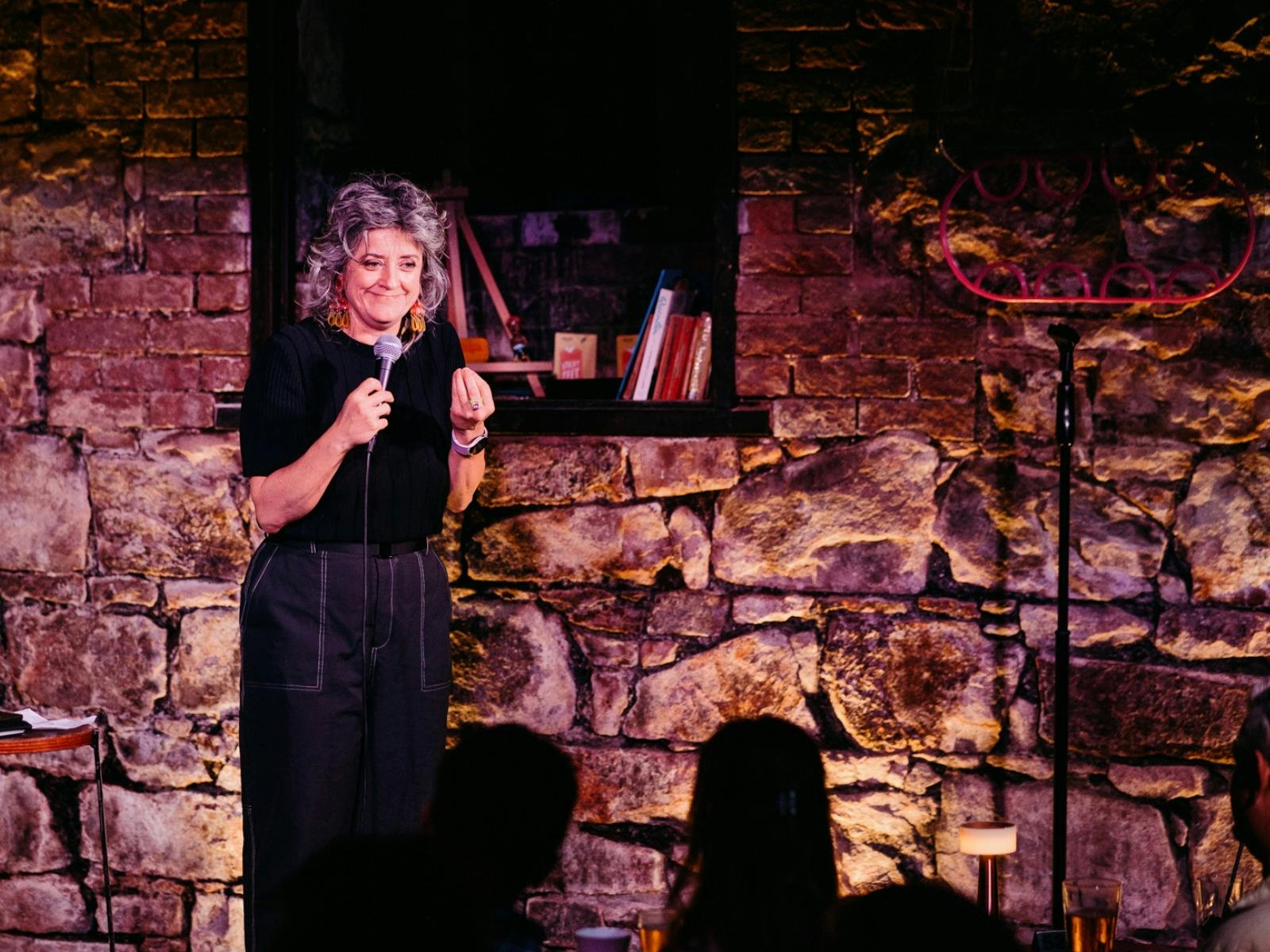 Comedian Chris Ryan on stage at a comedy club, with a heritage brick wall behind her. Comedian Chris Ryan on stage at a comedy club, with a heritage brick wall behind her.