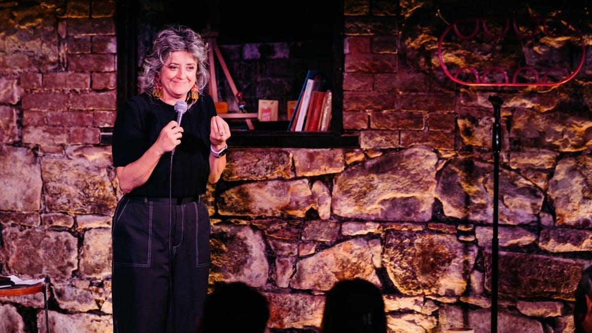 Comedian Chris Ryan on stage at a comedy club, with a heritage brick wall behind her.