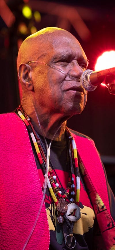 Uncle Archie roach sings on stage, wearing a black shirt and red waistcoat