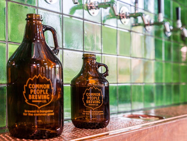 A large growler jug and smaller squealer jug sit on the copper bar under the green tiled tap wall