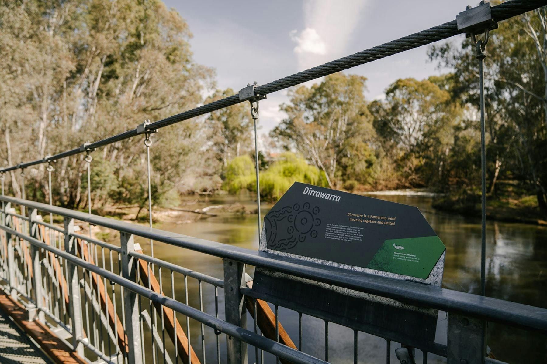 Edge of a swing bridge, over a river with a grey sign about cultural information.