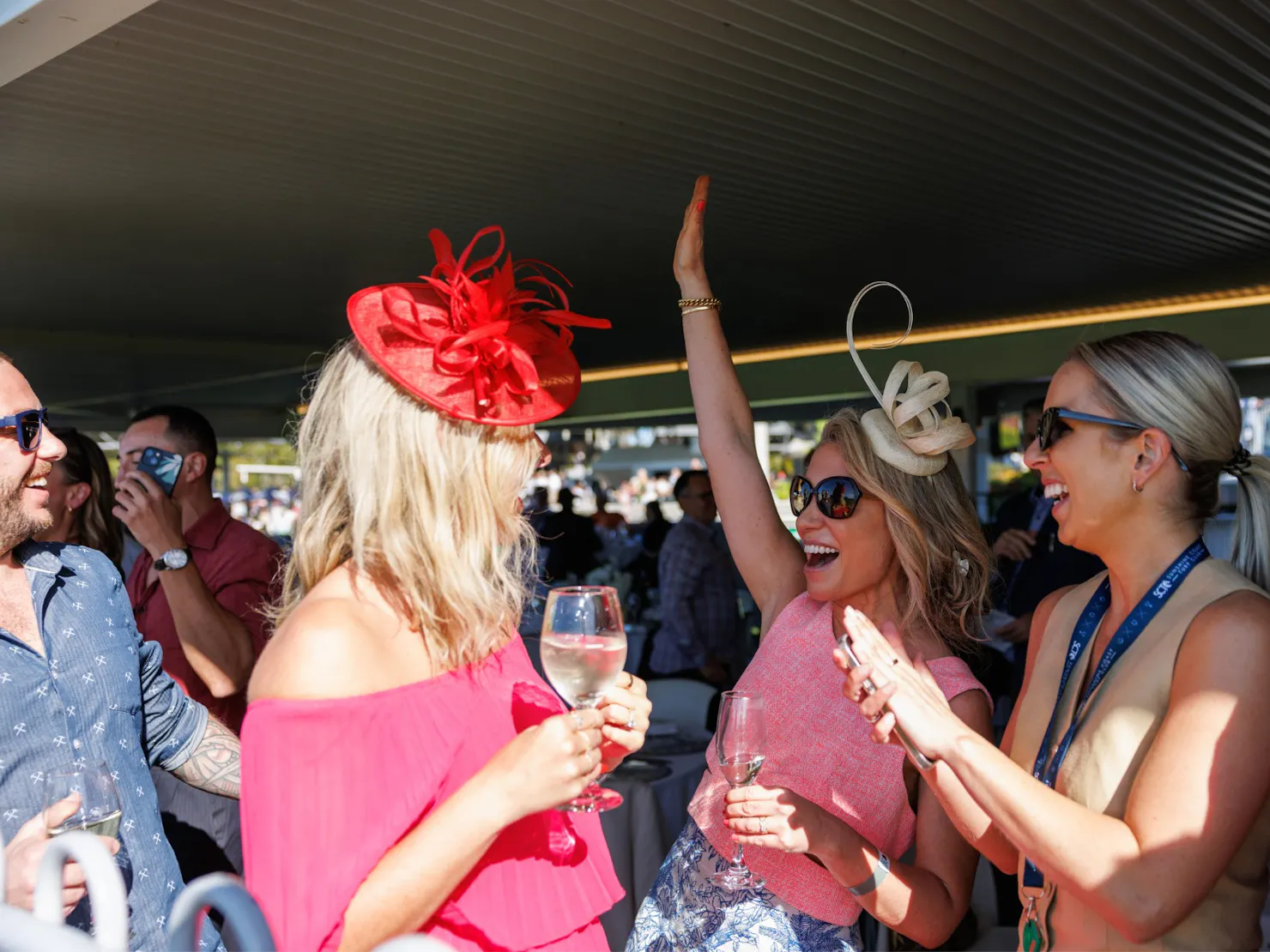 A group of women in racewear and fascinators laughing and high-fiving while holding glasses of wine,