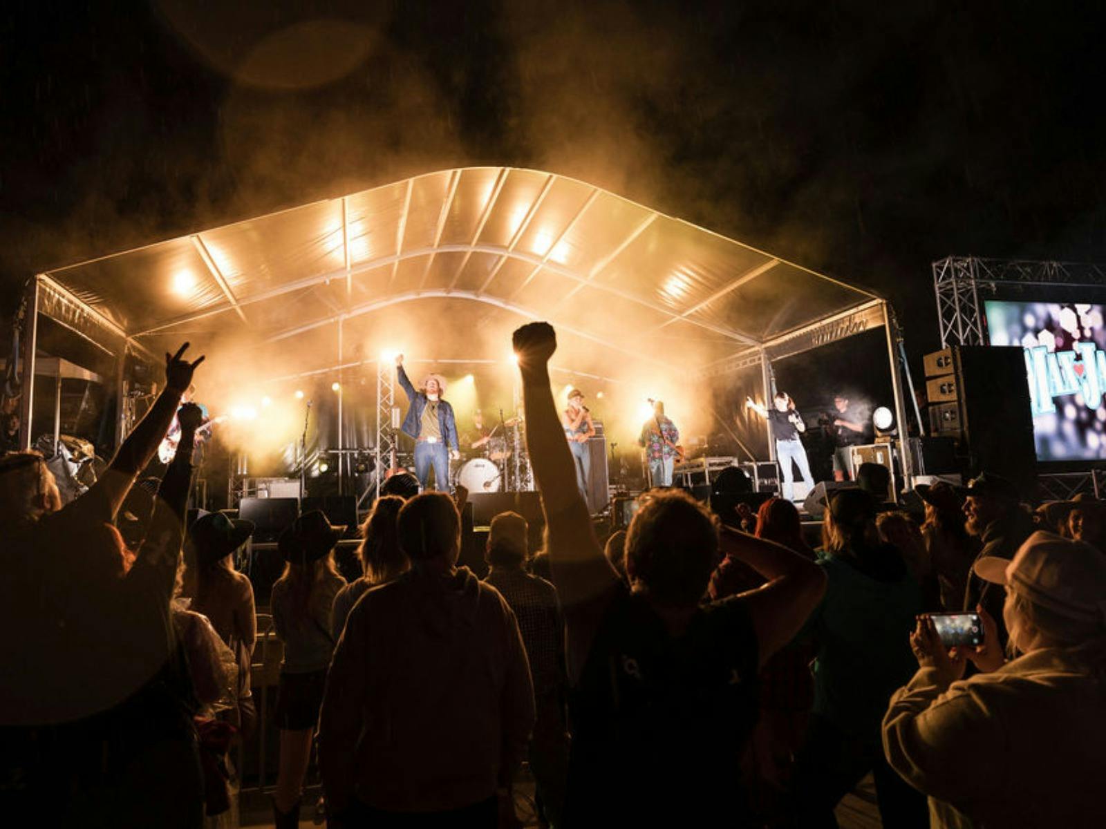 Audience watching a live outdoor concert at night with bright stage lights and raised hands.