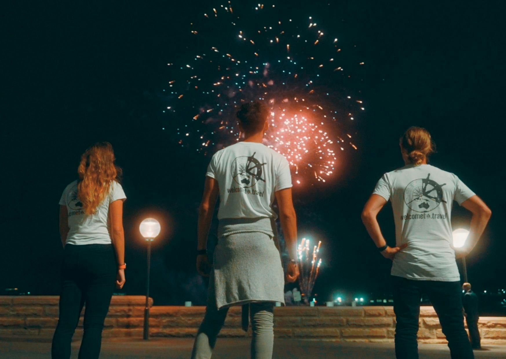 Three people looking up at fireworks with the Welcome to Travel logo on their shirts