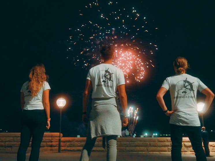 Three people looking up at fireworks with the Welcome to Travel logo on their shirts