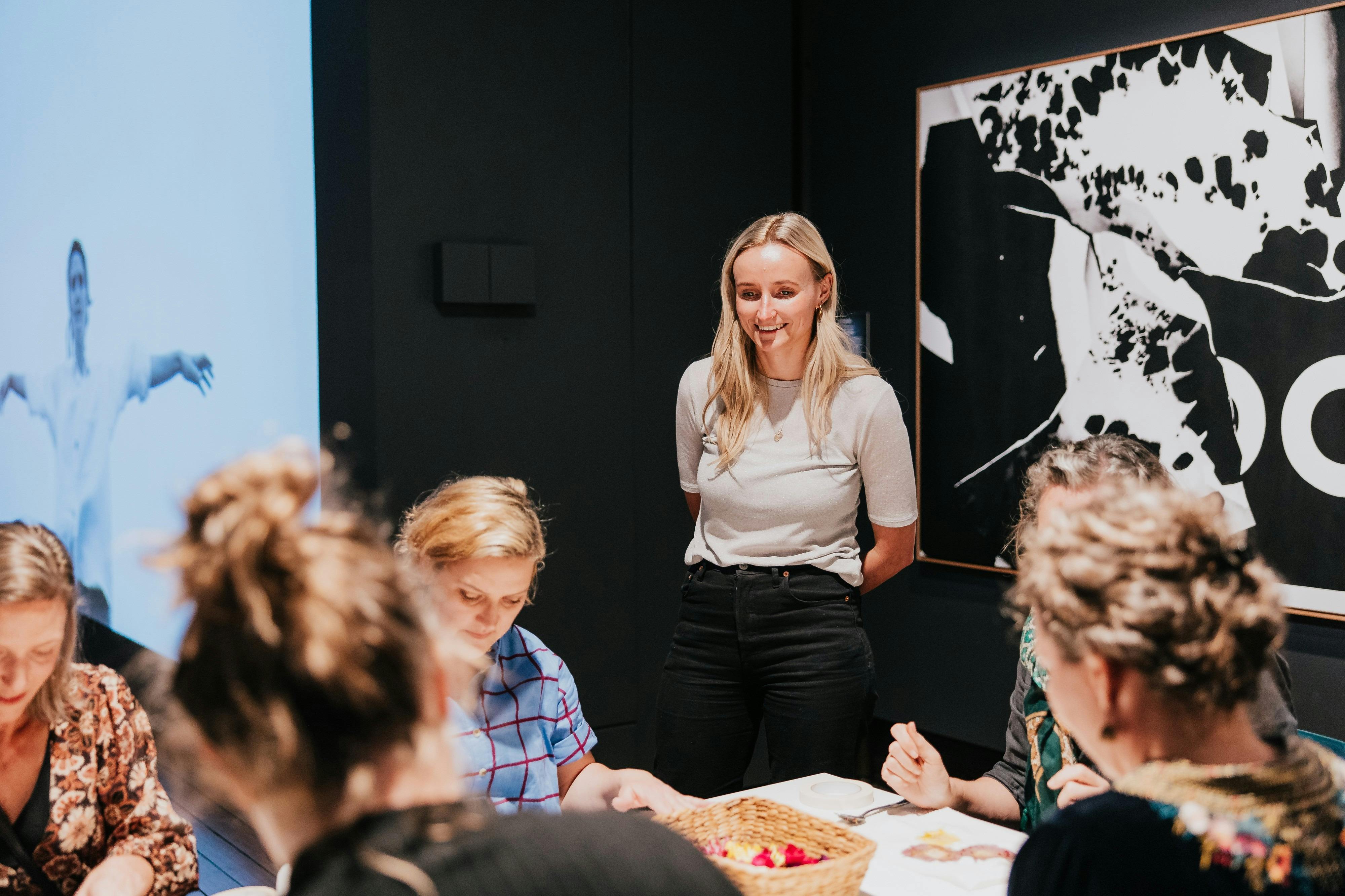 A blonde woman stands and watches women creating art