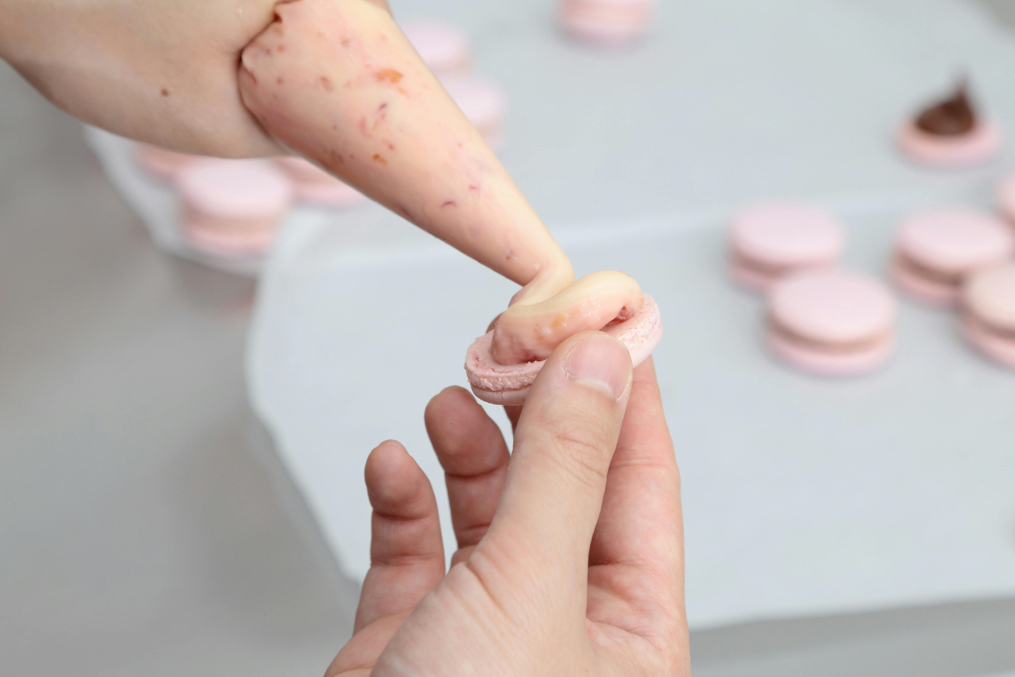 A hand piping pink filling onto a pink macaron shell in the Jacican cooking school kitchen Gippsland