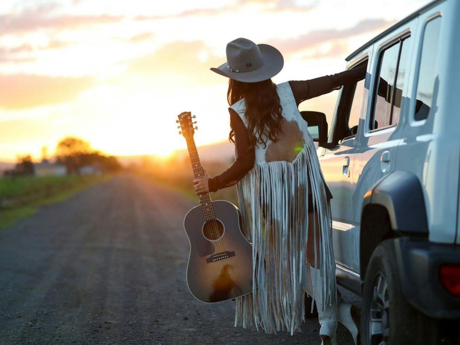 Tori Darke leaning away from white 4WD car holding her guatar and looking into the sunset