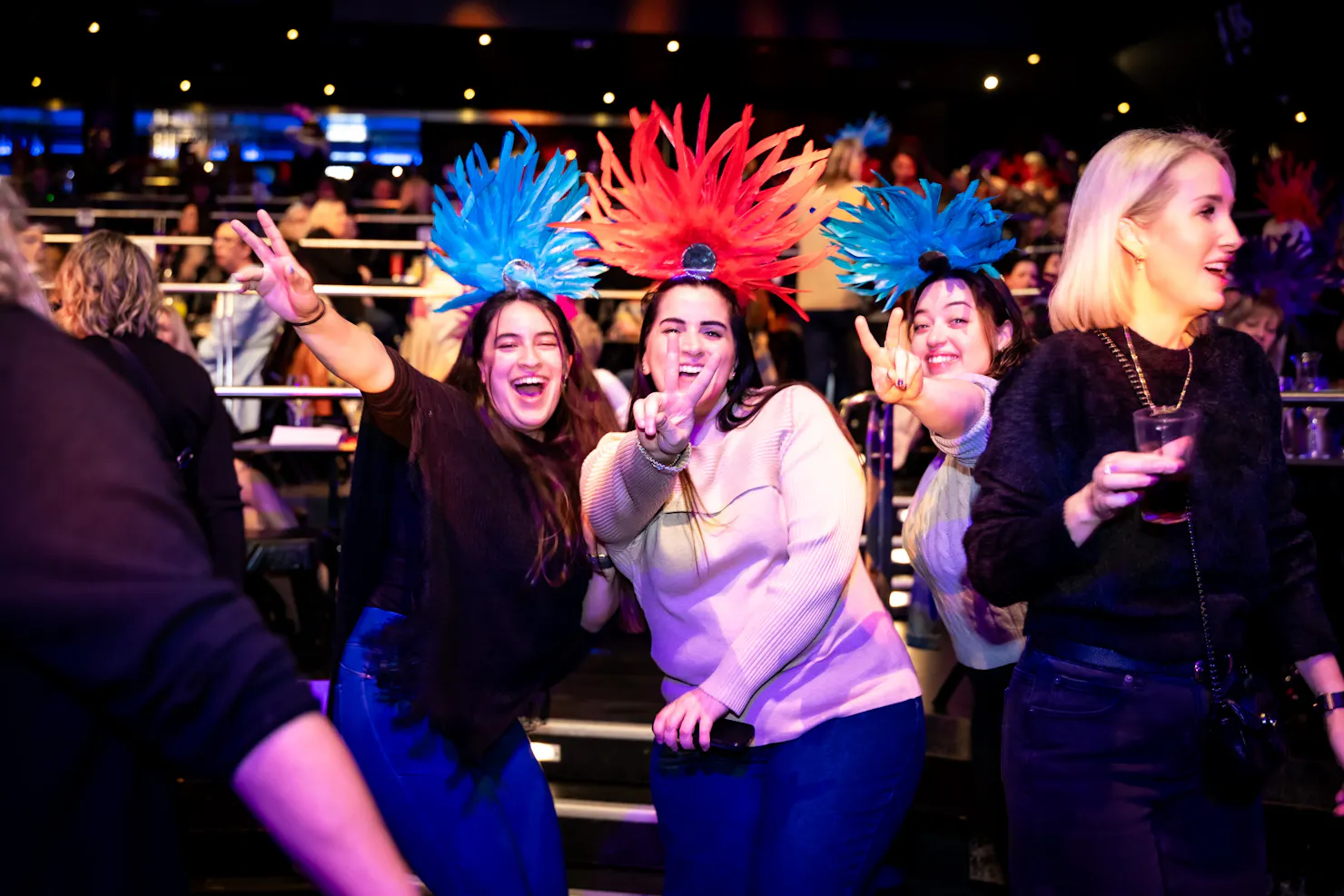 Three women wearing feathered headbands smile and give peace signs