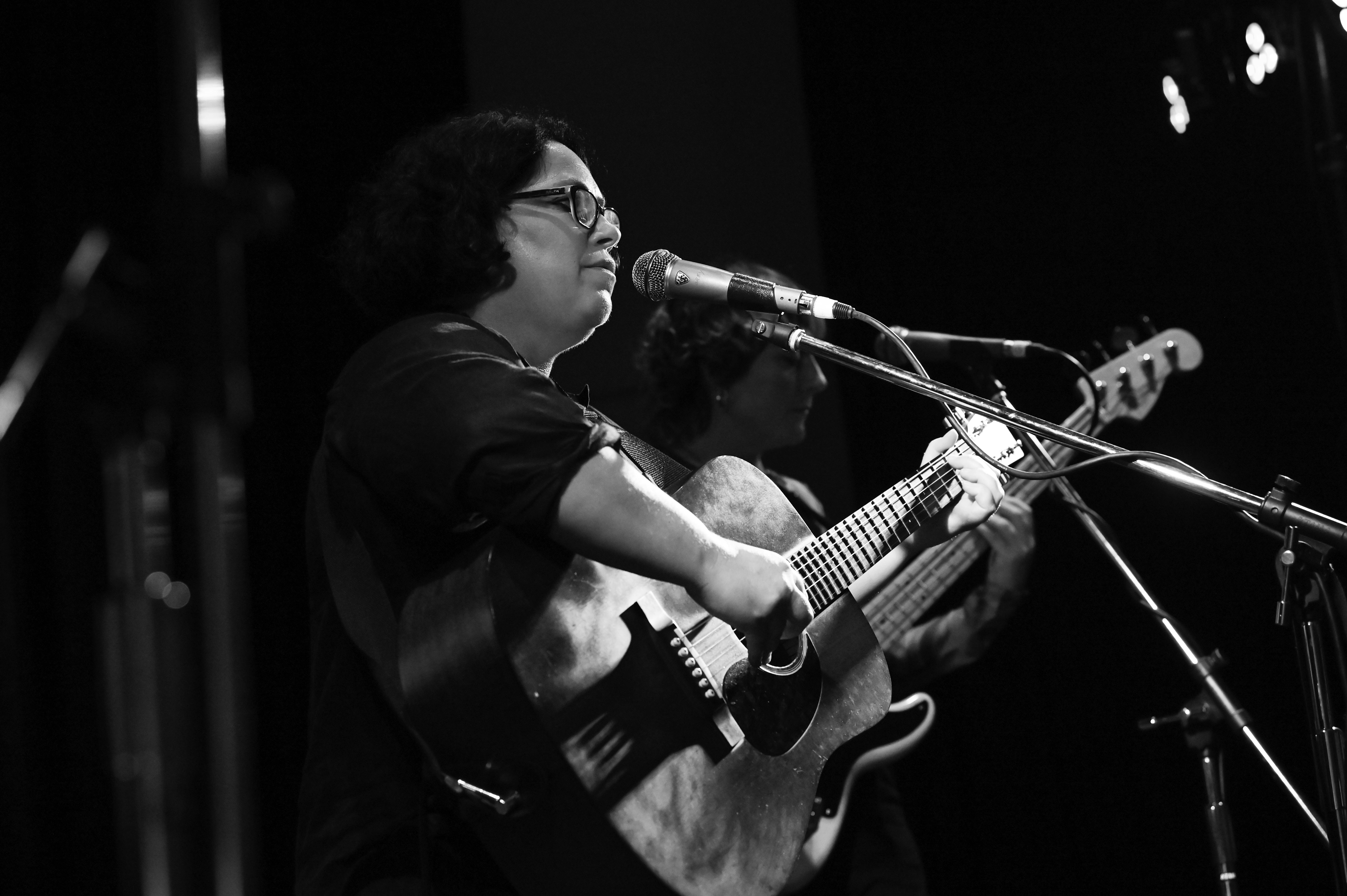 Nancy Bates holding a guitar in front of a microphone, behind her are other musicians.