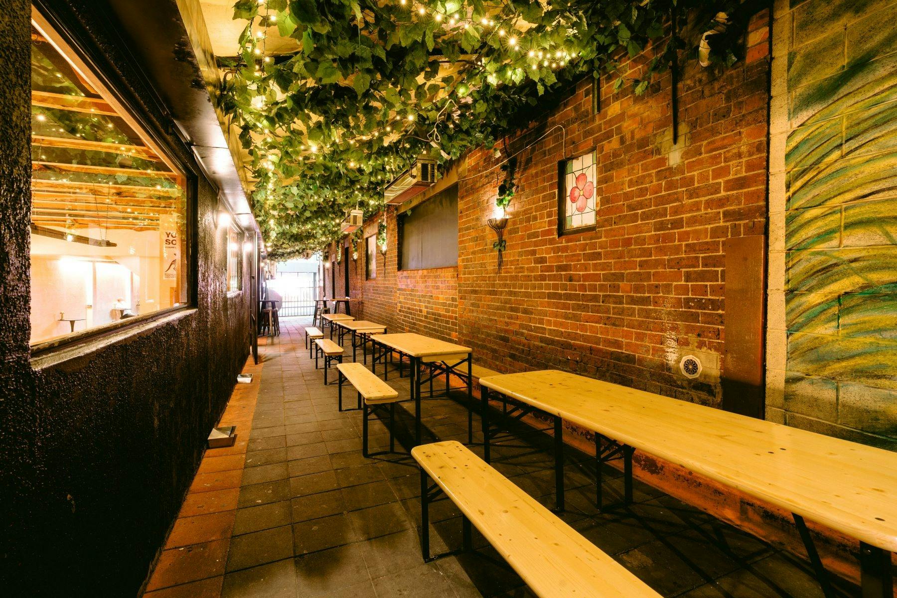 Outdoor beer garden with wooden tables, brick walls, and string lights under leafy canopy