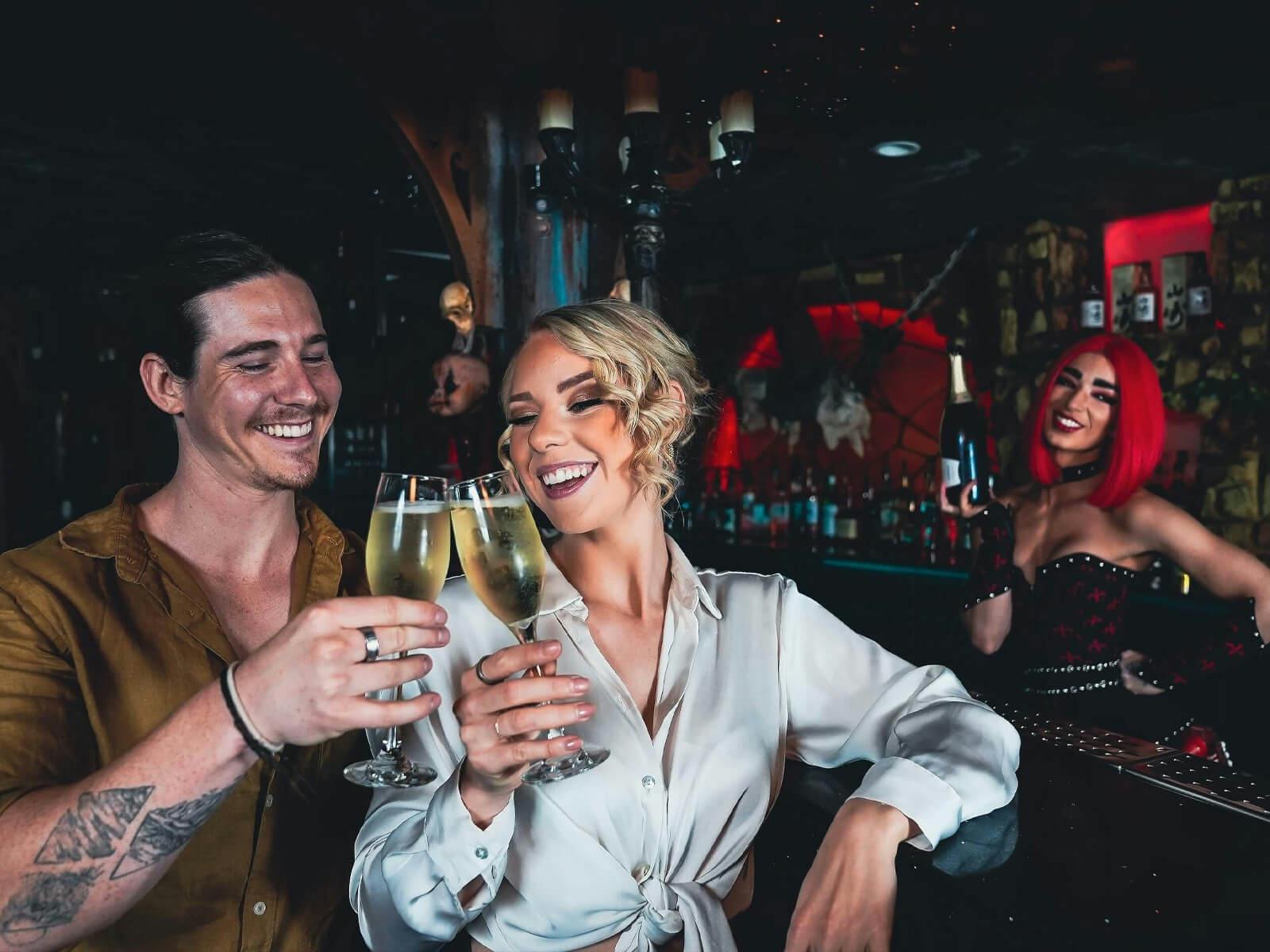 Two guests toast with sparkling wine at the Dracula's bar while a bartender in costume smiles behind