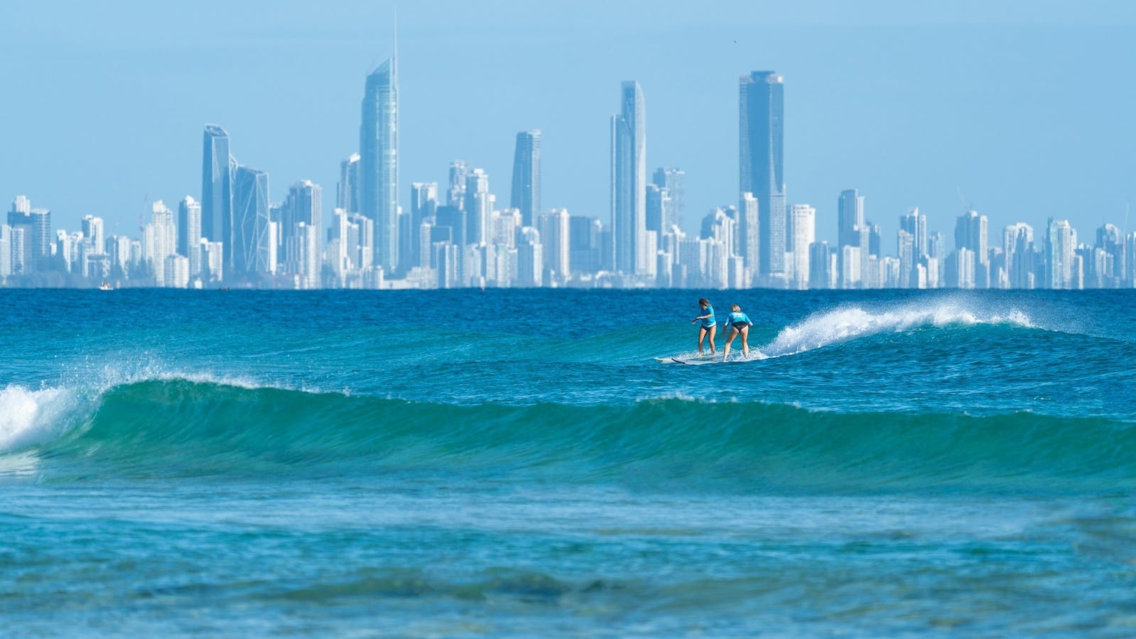 Two women surfing in blue ocean with city skyline in background. Two women surfing in blue ocean with city skyline in background.