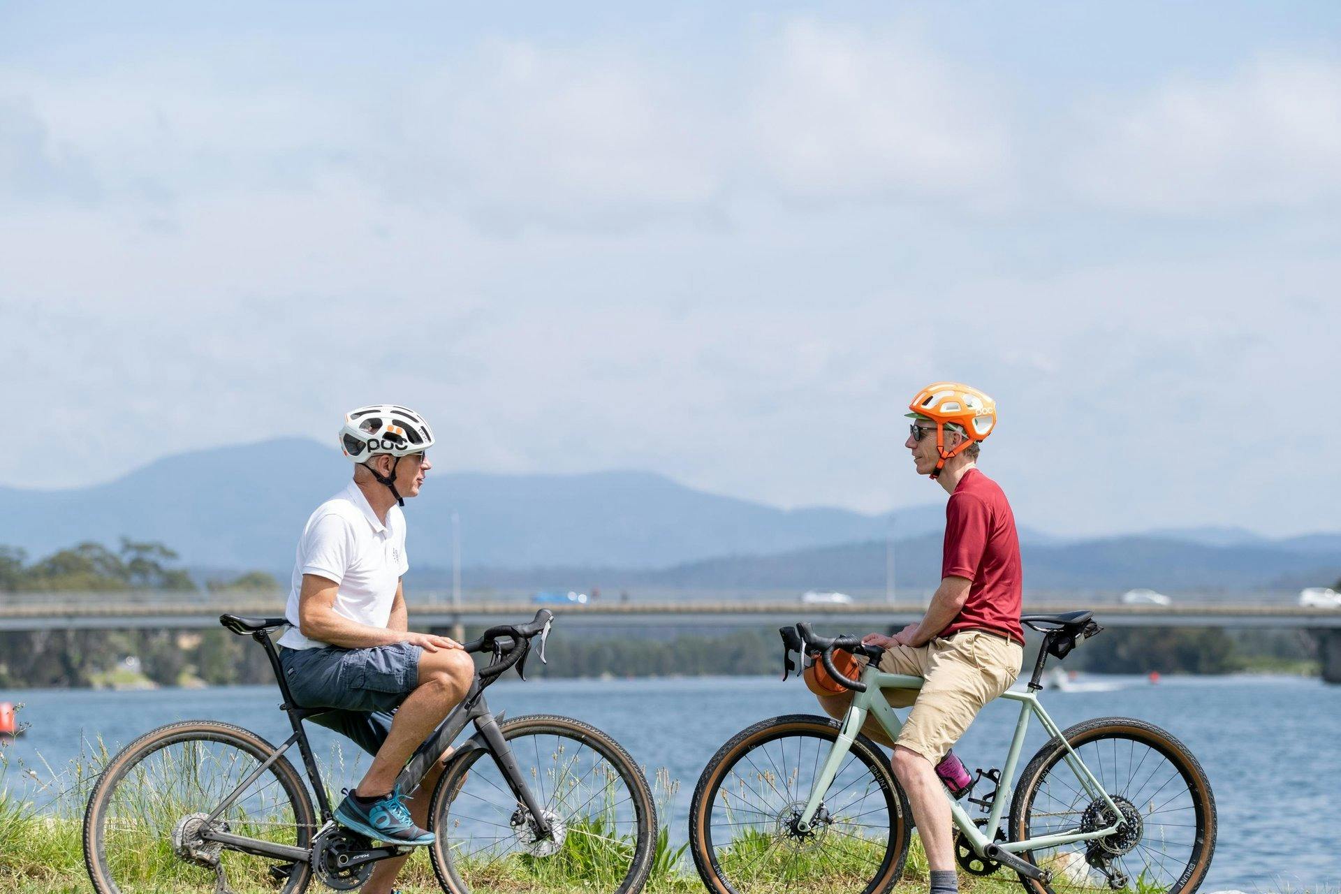 Two cyclists facing each other with hte Moruya River and mountains in the background