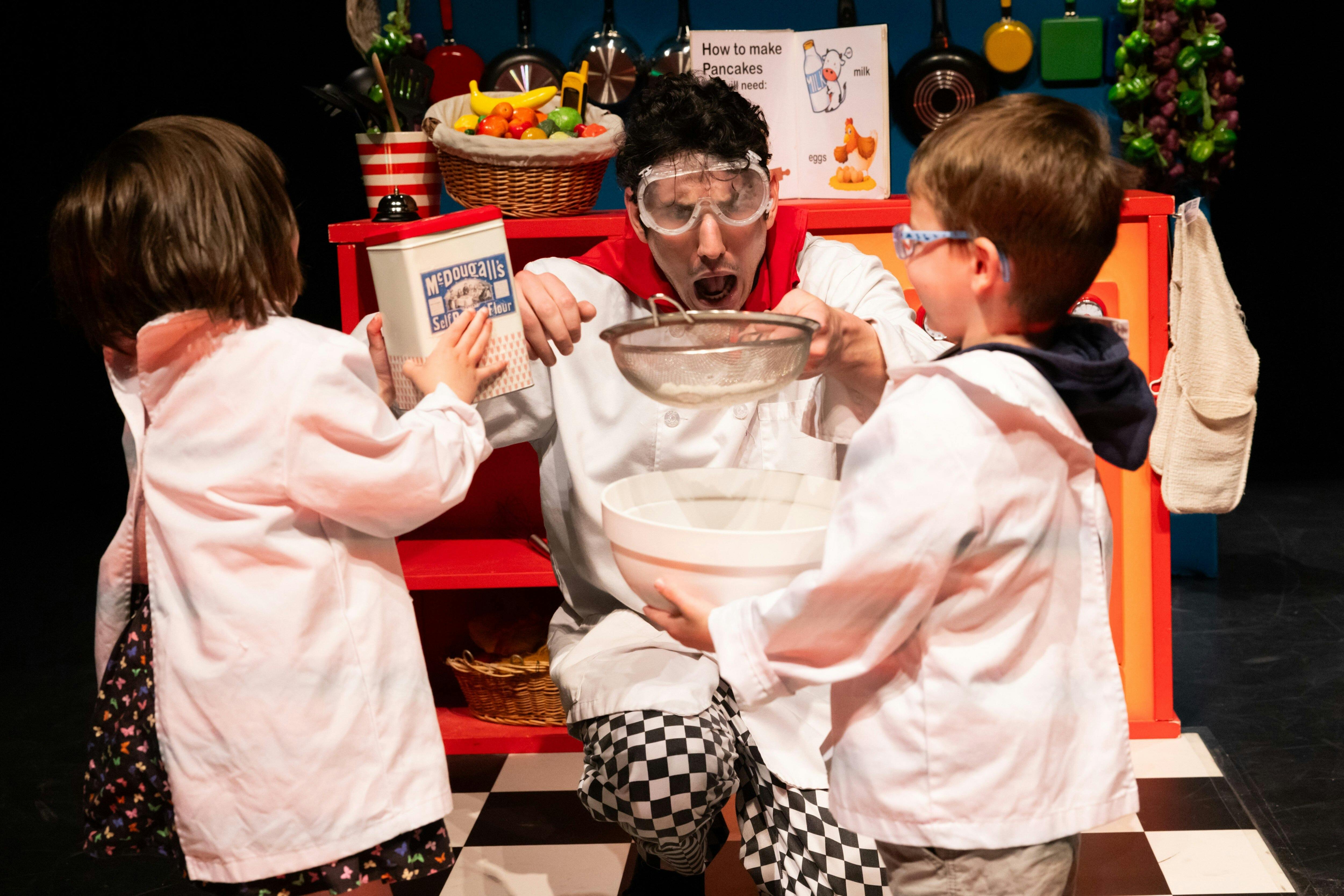 A person in goggles, holding a sieve over a mixing bowl, looks surprised in a playful kitchen scene