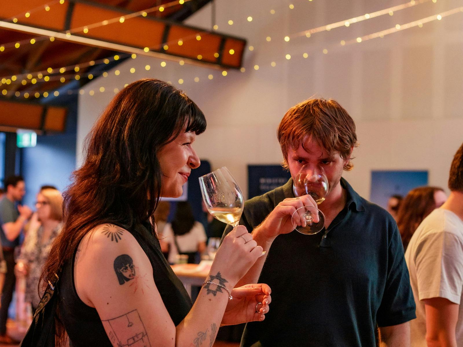 A woman swirls her glass beside a man smelling his; warm string lights in the background