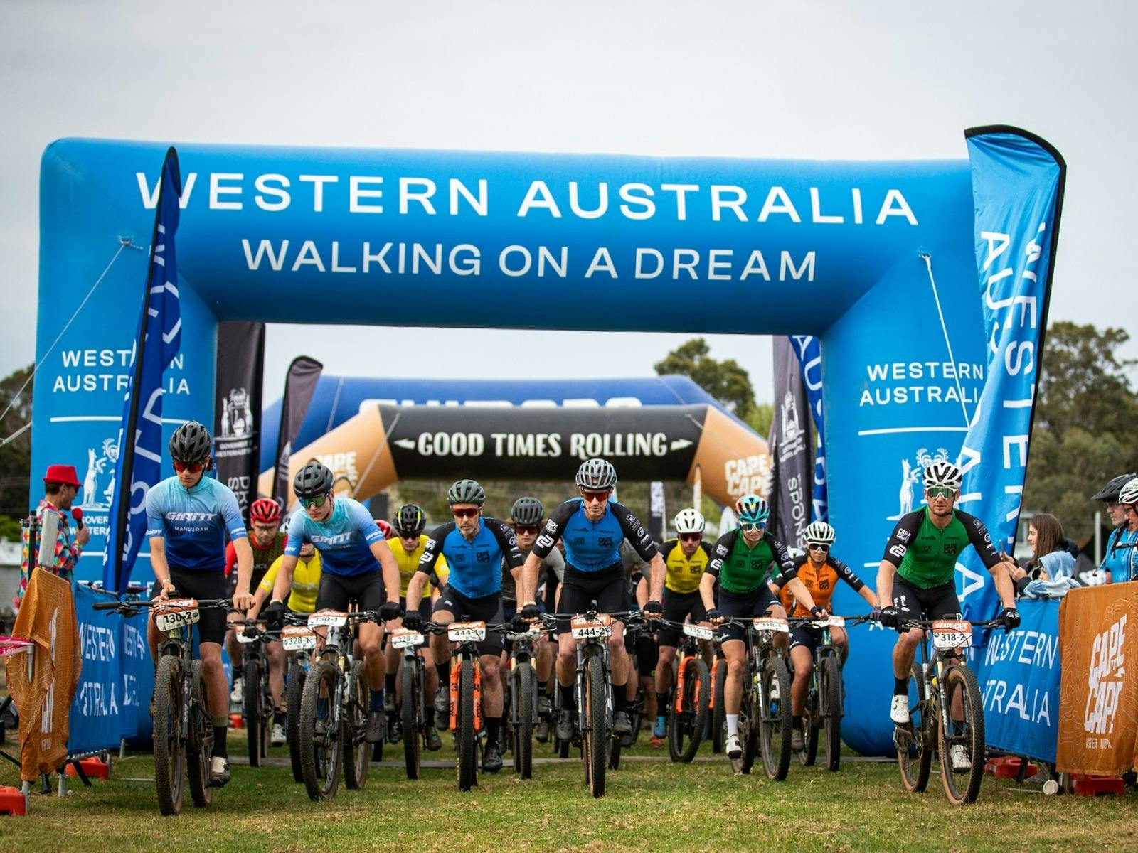 Group of mountain bike riders starting the event, riding under blue Tourism WA arch and signage